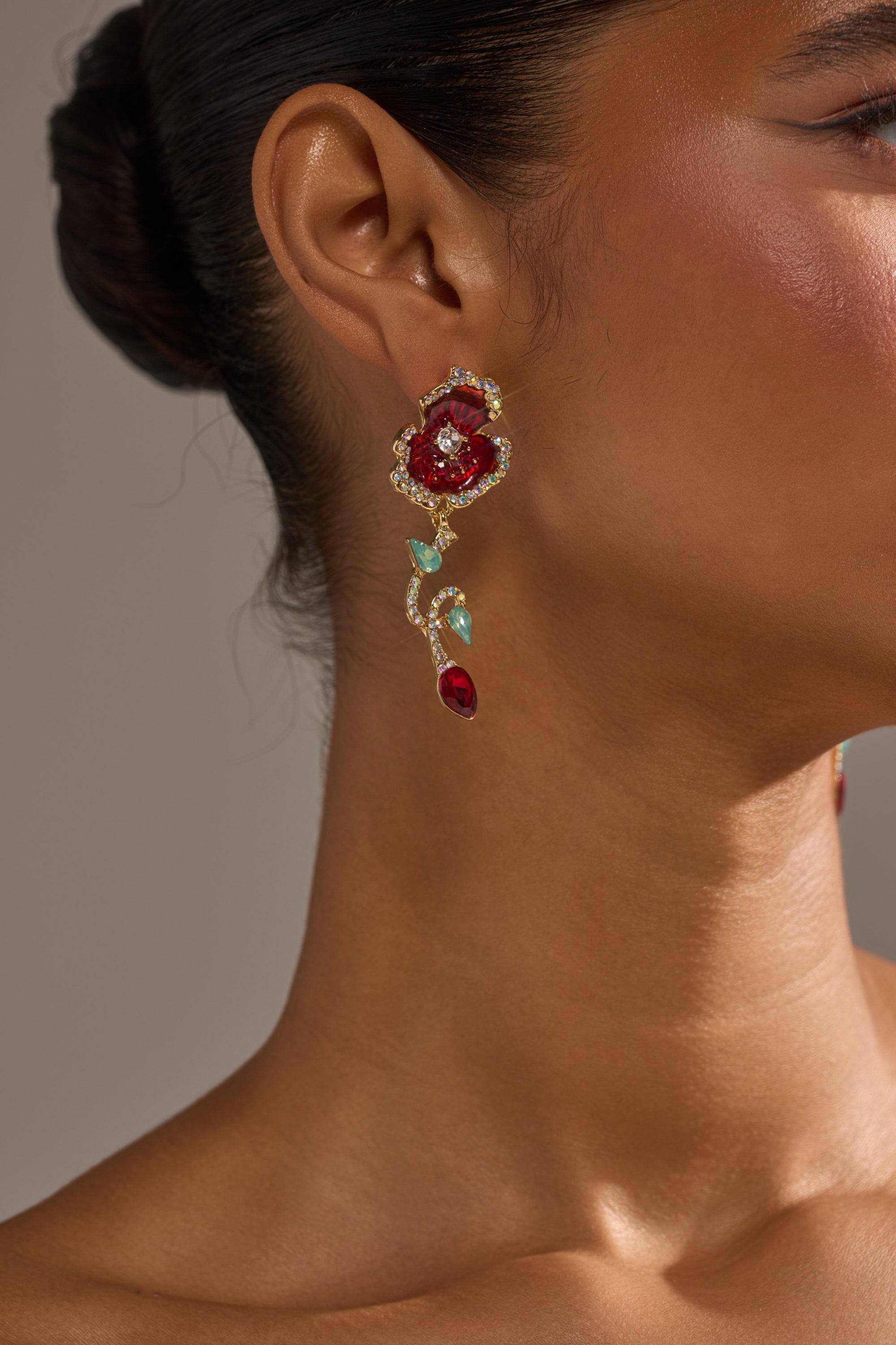 A close-up of a woman's ear adorned with the ROSEY EARRING, an ornate floral-shaped accessory with red, turquoise, and gold details. She has a smooth complexion and dark hair styled in a neat bun. 1
