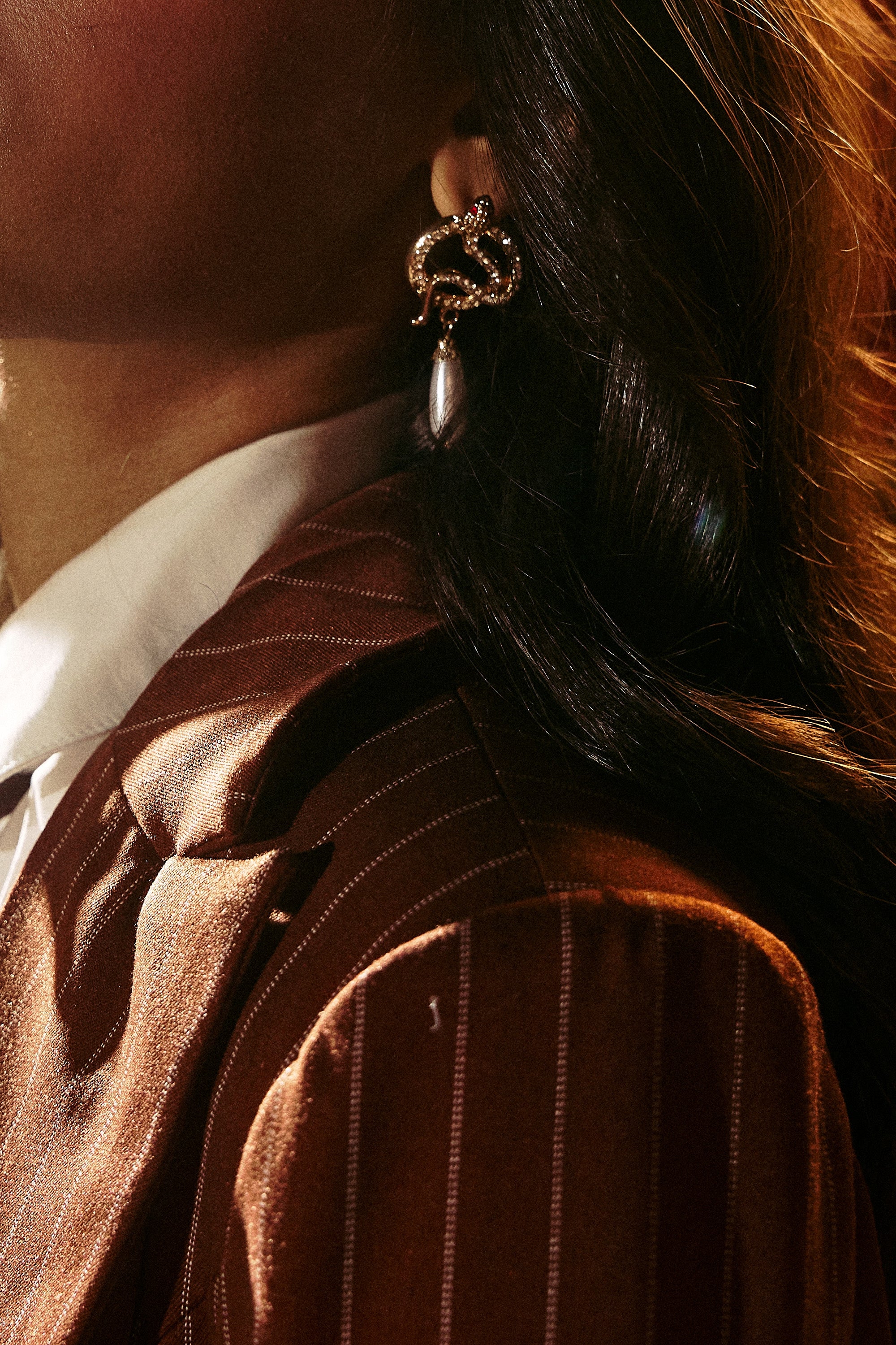 Close-up of a person in a brown pinstriped blazer and white collared shirt, with wavy hair wearing the UR A SNAKE EARRING, a decorative gold earring featuring a dangling pearl accent.
