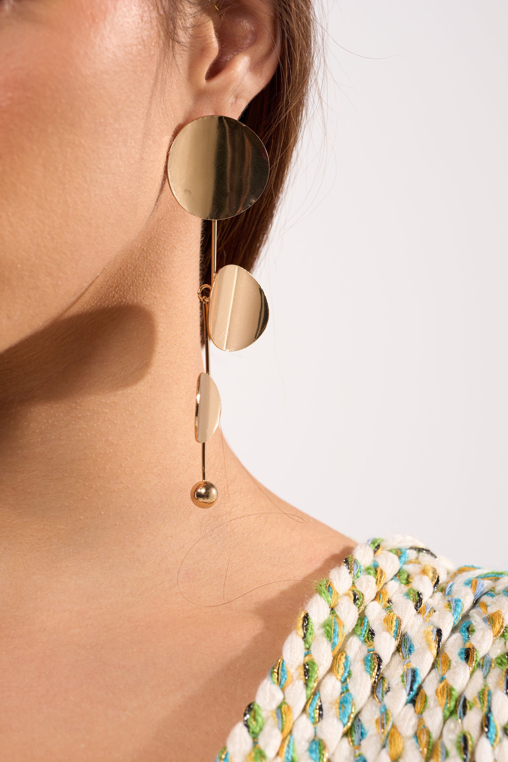 Close-up of a person wearing the DEDE EARRING—large, modern gold drop earrings with geometric circles—paired with a textured, multicolored woven top. Only part of the face, ear, neck, and shoulder are visible. 1