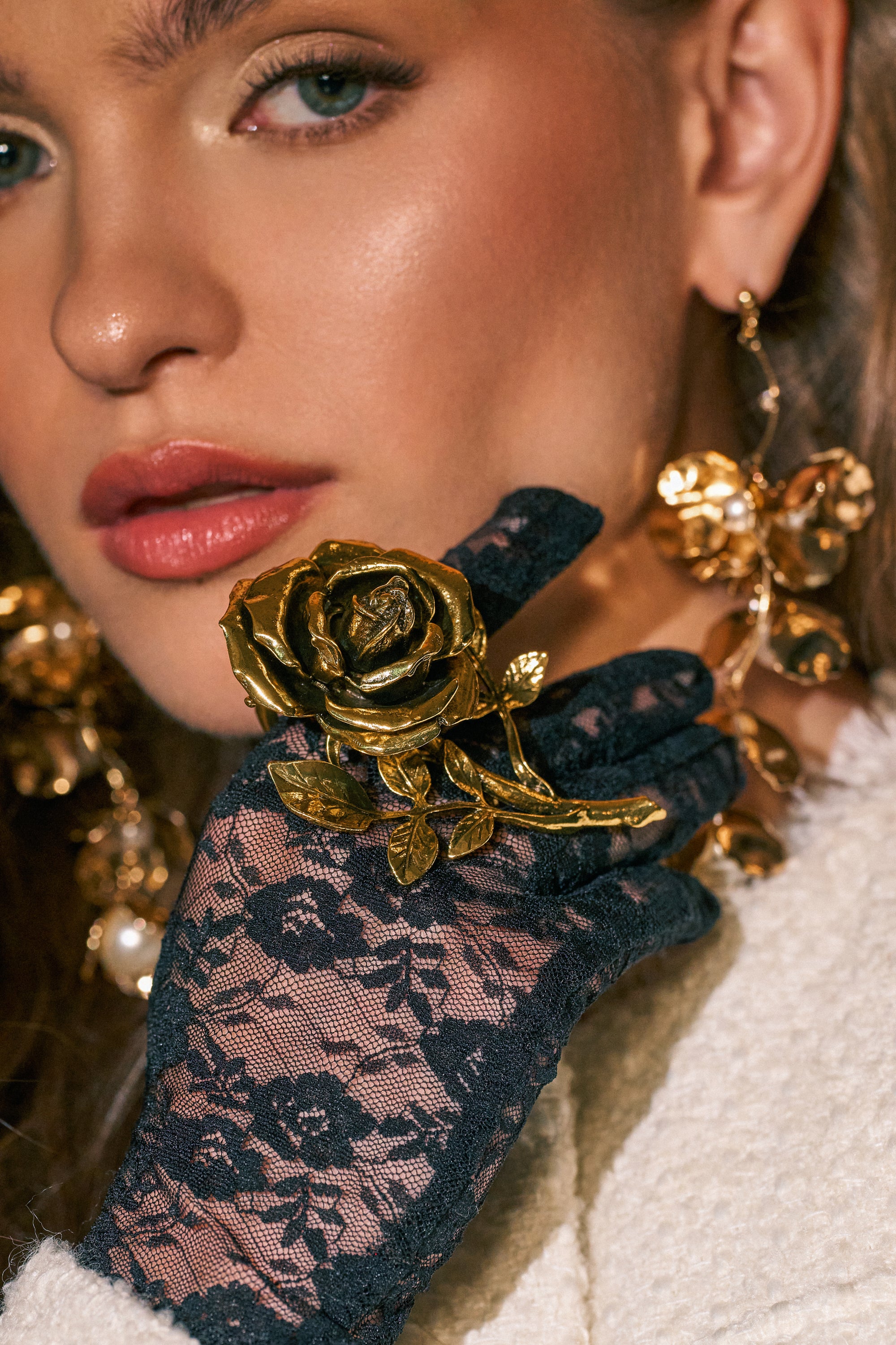 A woman wearing black lace gloves holds the IN BLOOM COCKTAIL RING—a large gold-tone rose design—near her face. She pairs it with gold floral earrings and a white textured garment. Her soft, glowy makeup emphasizes her eyes and lips, drawing attention to the striking statement ring she displays with elegance.