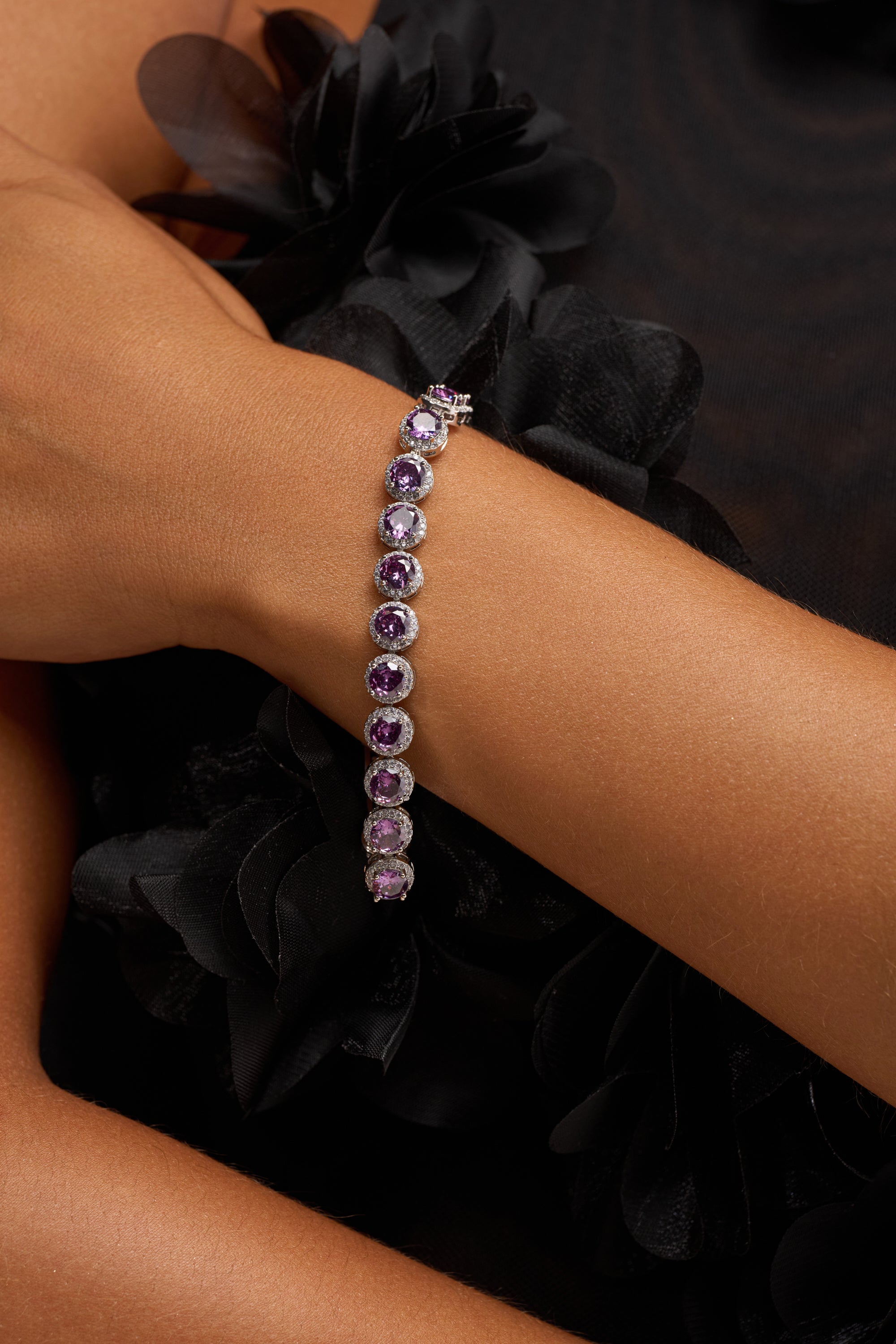 A close-up of a person’s arm wearing the PLUM PLS CZ BRACELET, featuring round plum gemstones on a silver-tone band, paired with a black outfit detailed with fabric flowers—a sophisticated formalwear accessory.