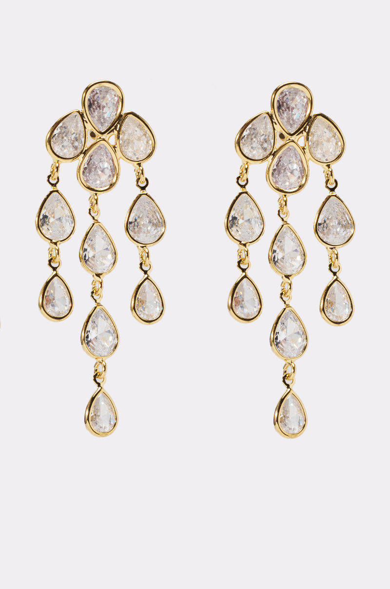 The LOVE ME EARRING showcases gold-tone drop chandelier earrings with clusters of teardrop-shaped clear gemstones in three cascading rows, displayed against a white background. 0