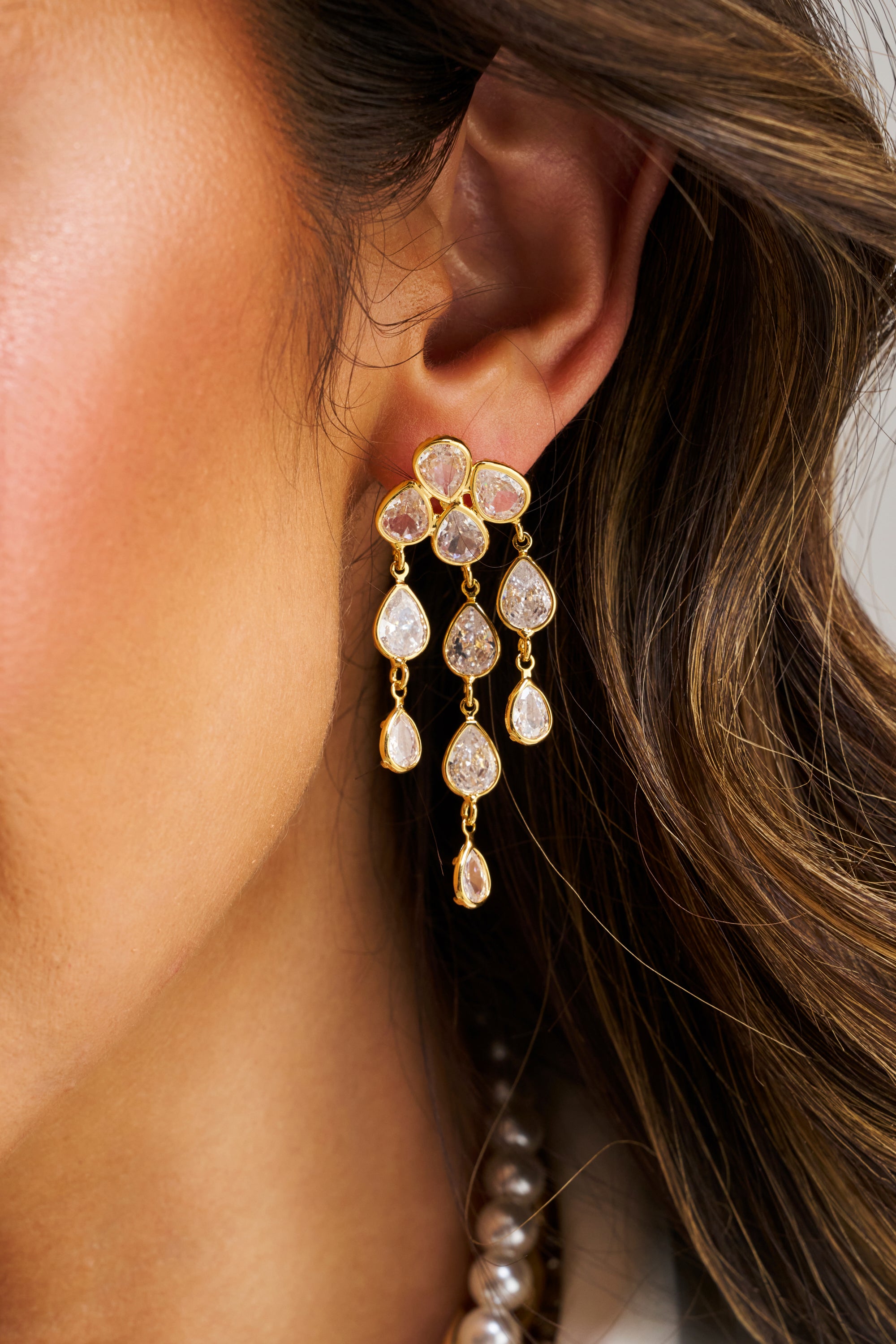 Close-up of a woman's ear wearing LOVE ME EARRING—gold-tone drop earrings featuring multiple teardrop-shaped clear gemstones. Her wavy brown hair and part of a pearl necklace are also shown.