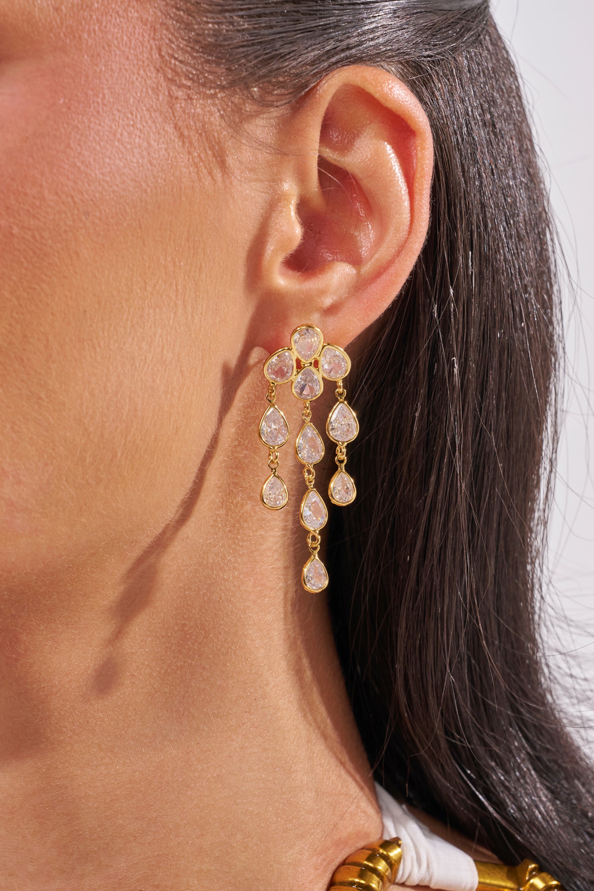 Close-up of a woman's ear wearing the LOVE ME EARRING—elegant gold drop earrings with multiple teardrop-shaped clear gemstones, reminiscent of chandelier style, seen against straight dark hair and light skin. 1