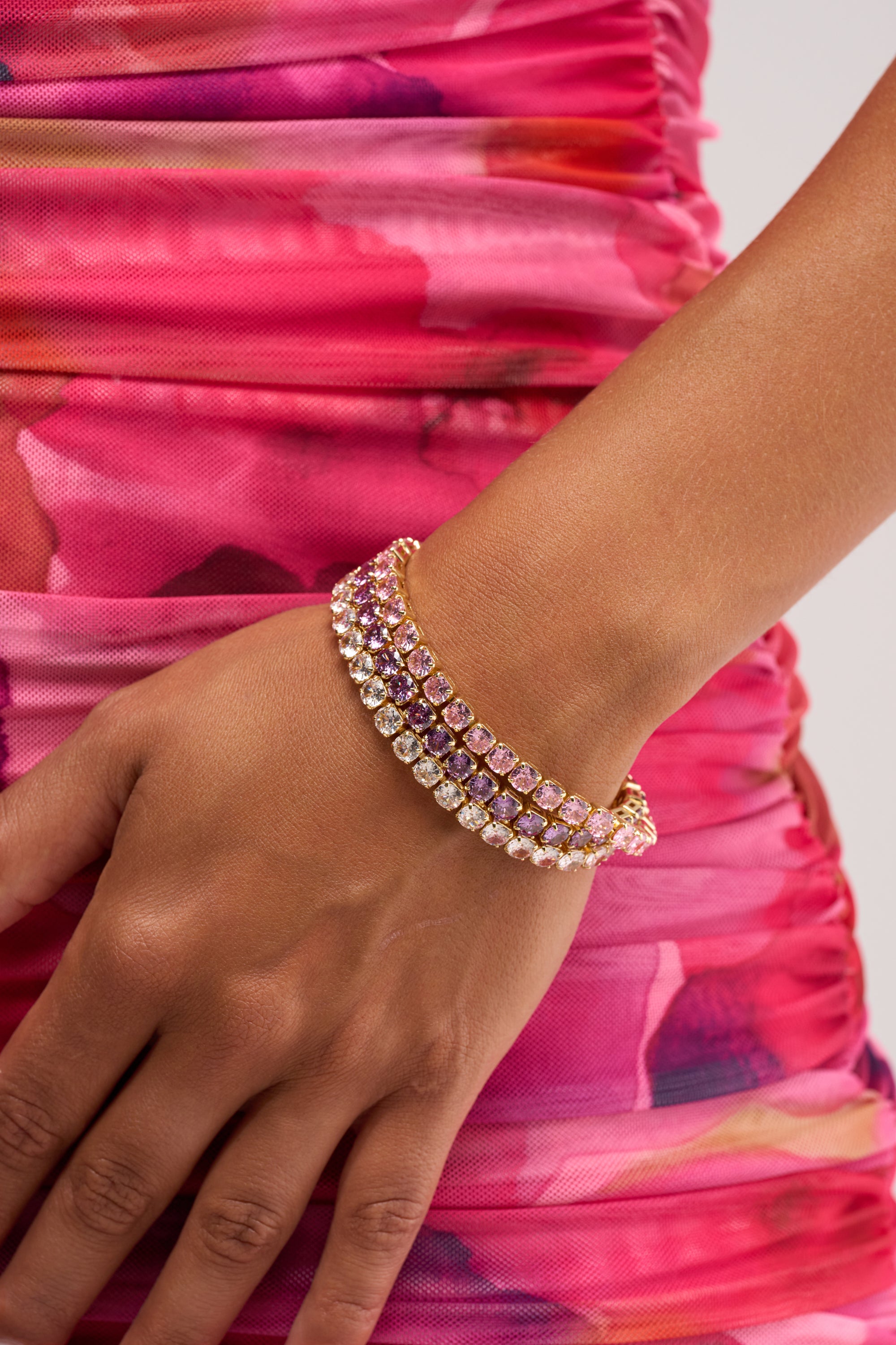 A close-up of a hand wearing the IT GIRL BRACELET SET, featuring gold-tone bracelets with purple and clear gemstones, resting against a pink and red floral-patterned dress.