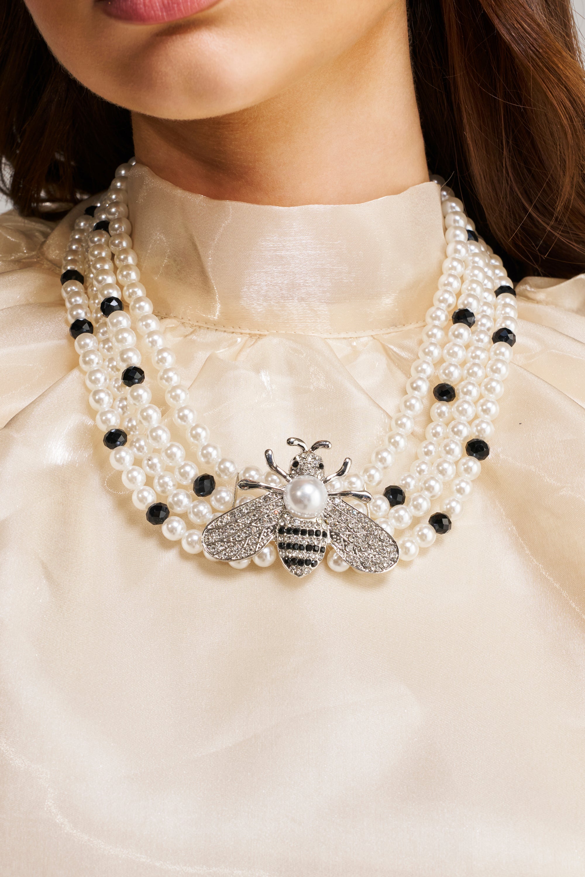 A woman wearing a cream satin blouse and the QUEEN BEE NECKLACE, featuring pearls, black beads, and a large jeweled bee pendant at the center.