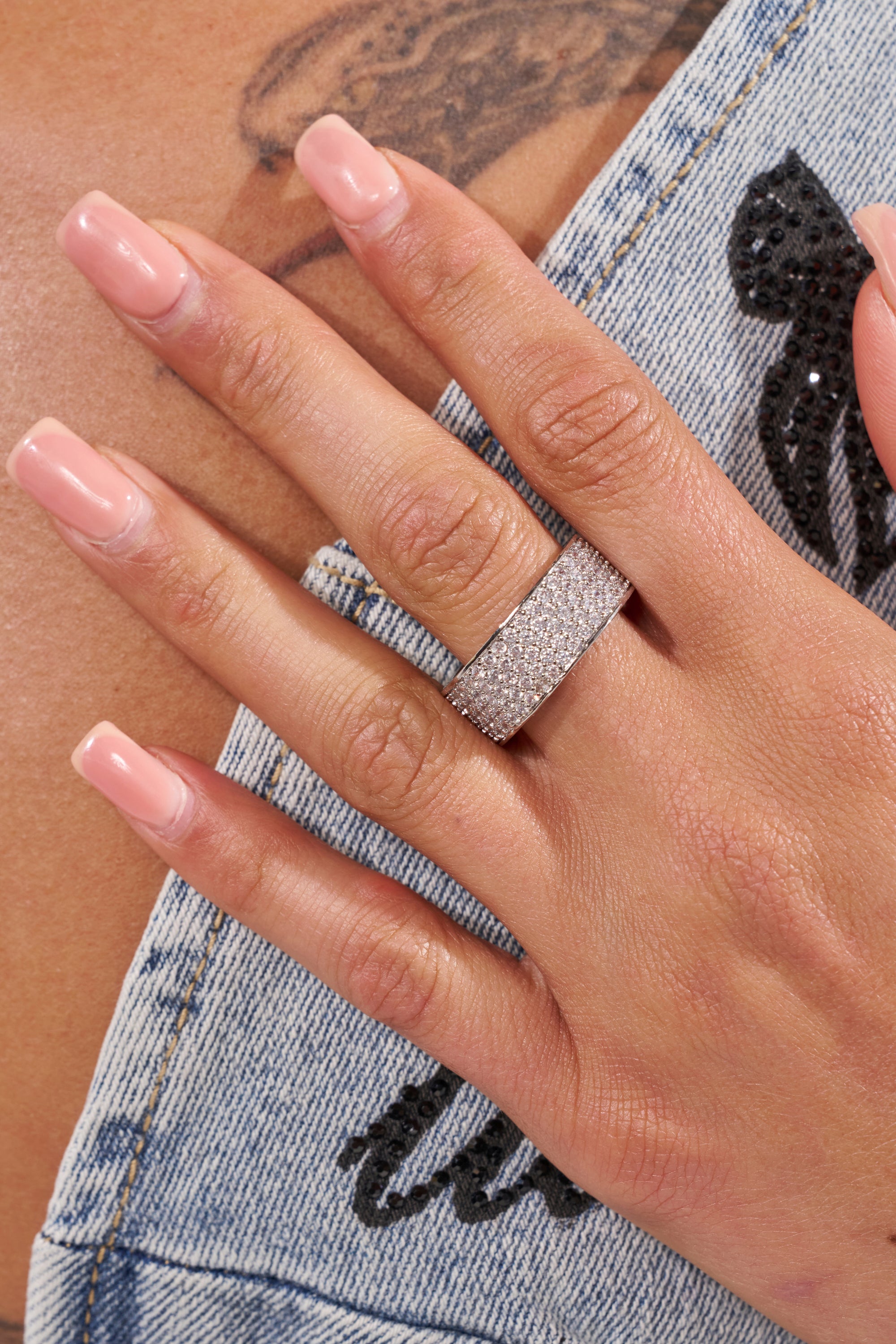 A hand with long, light pink manicured nails wears the TO INFINITY CZ RING, a wide rhinestone band. The hand rests on a tattooed shoulder covered by a blue denim jacket with black embroidery. 1