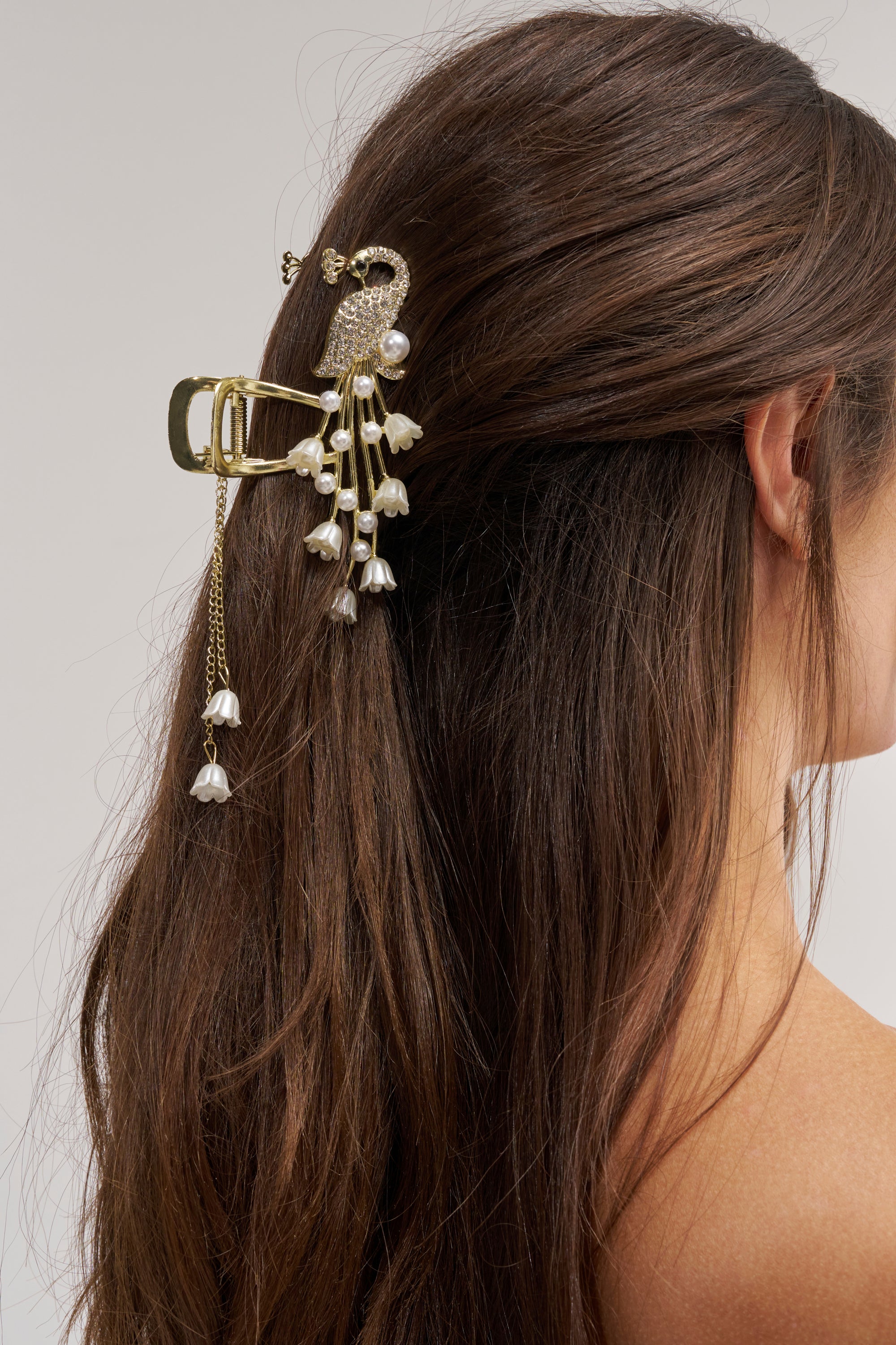 A woman with long brown hair wears the TAKE ME OUT HAIR CLAW CLIP, a decorative peacock-shaped accessory adorned with dangling pearls and delicate charms, against a plain background.