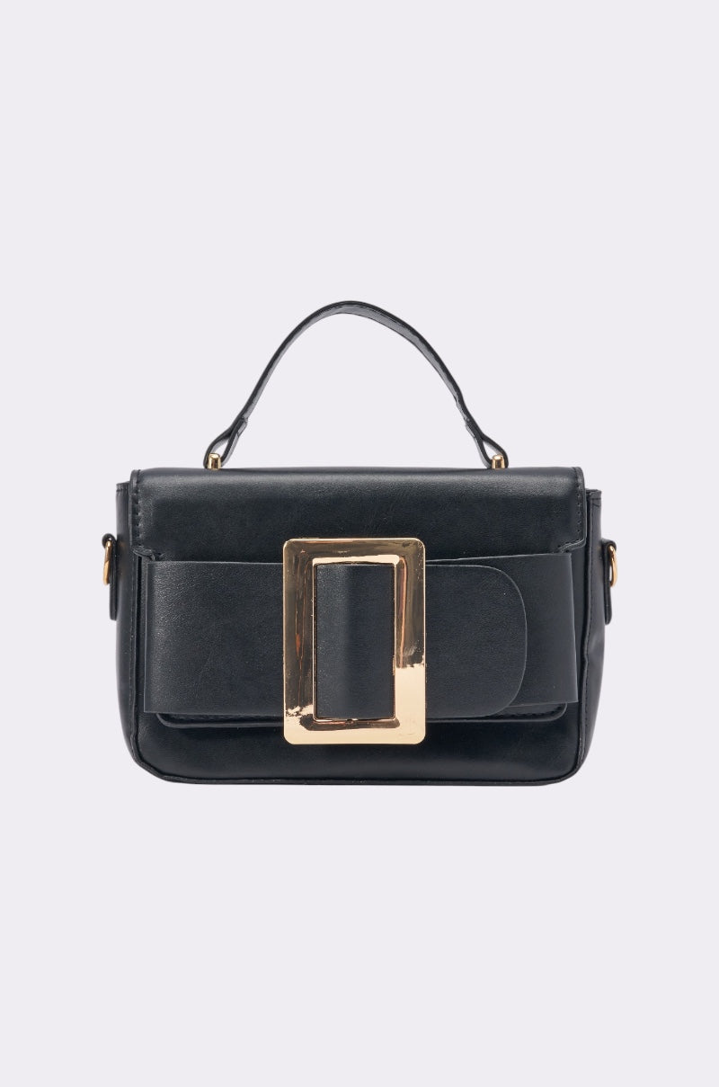 The DRESS ME UP PURSE is a small, structured black faux leather bag featuring a top handle and an eye-catching rectangular gold buckle on the front flap, displayed on a light gray background.