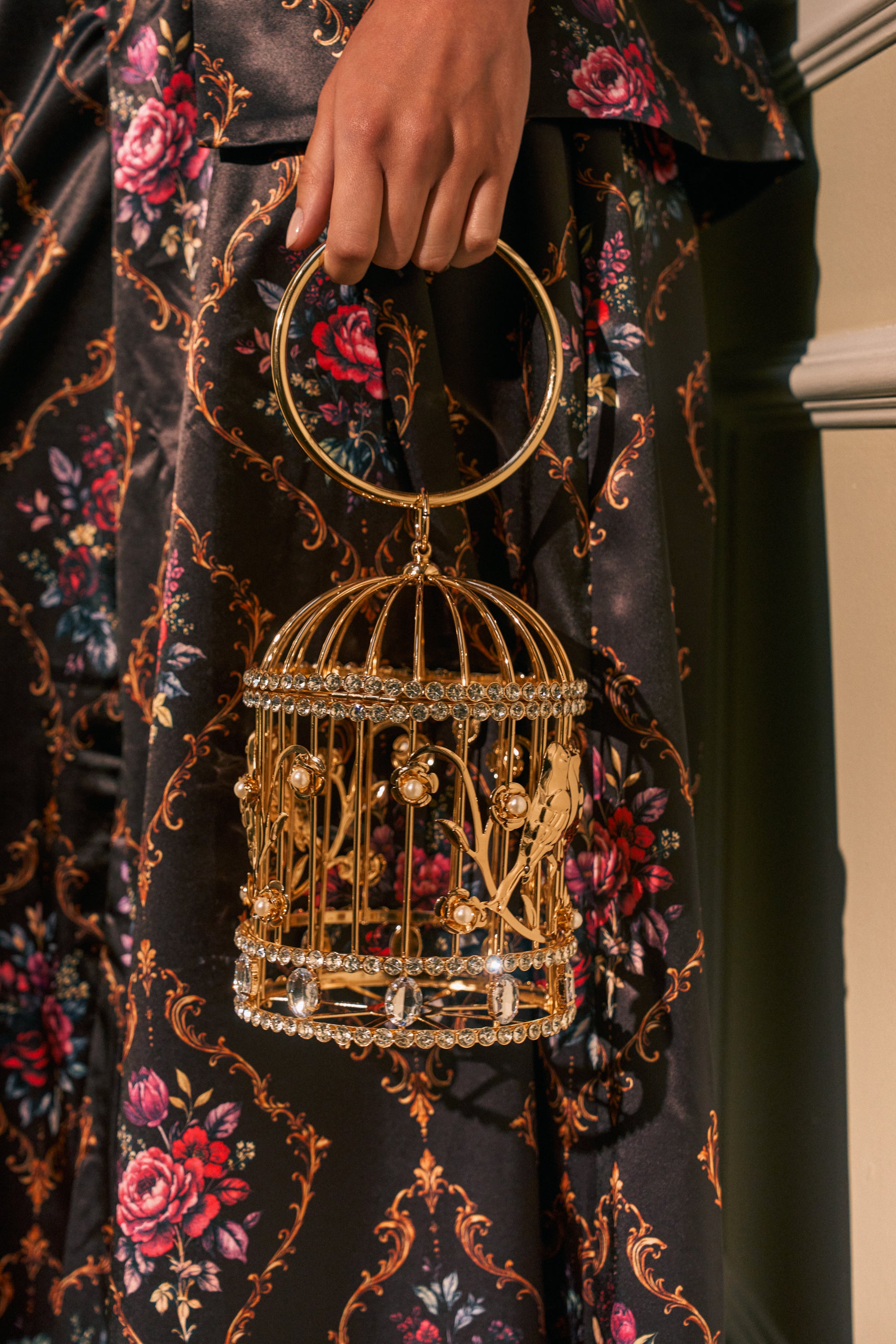 A hand holds the NEVER CAGED LUXE BAG, a decorative gold bird cage bag adorned with crystals, set against a floral-patterned satin dress.