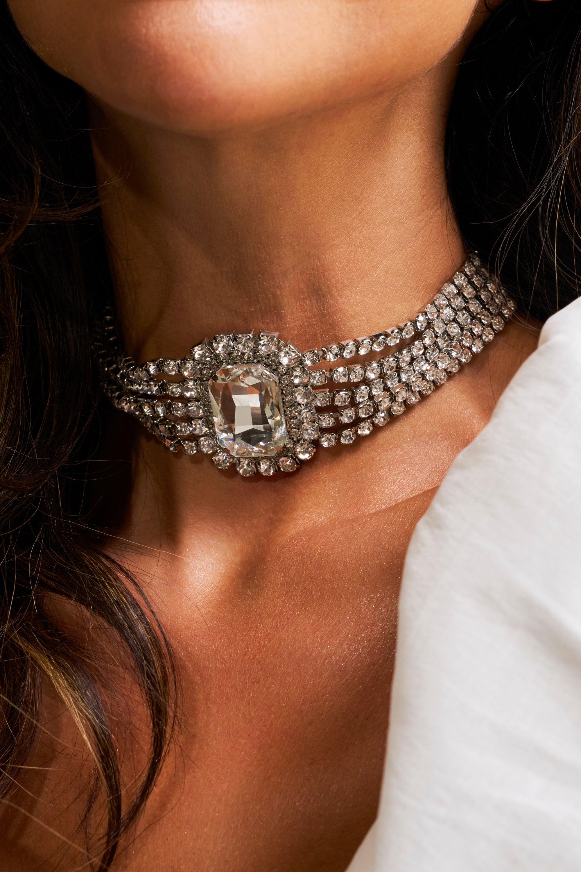 A close-up of a woman with wavy brown hair in a white top wearing the REGAL NECKLACE—a layered statement piece featuring a multi-strand diamond choker with a large rectangular gem at the center.