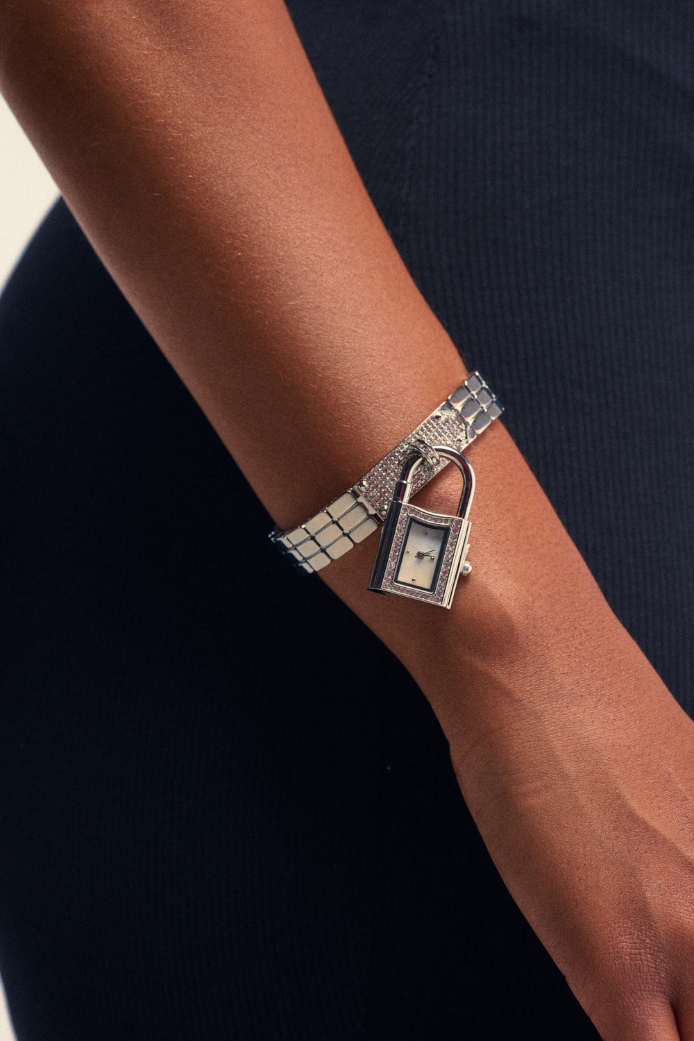 A close-up of a person’s wrist wearing the I LIKE KASH WATCH, featuring a distinctive padlock-shaped silver face set against a dark, textured sleeve. The watch boasts a small rectangular dial and a shiny, patterned strap, making it the perfect statement accessory for any occasion.
