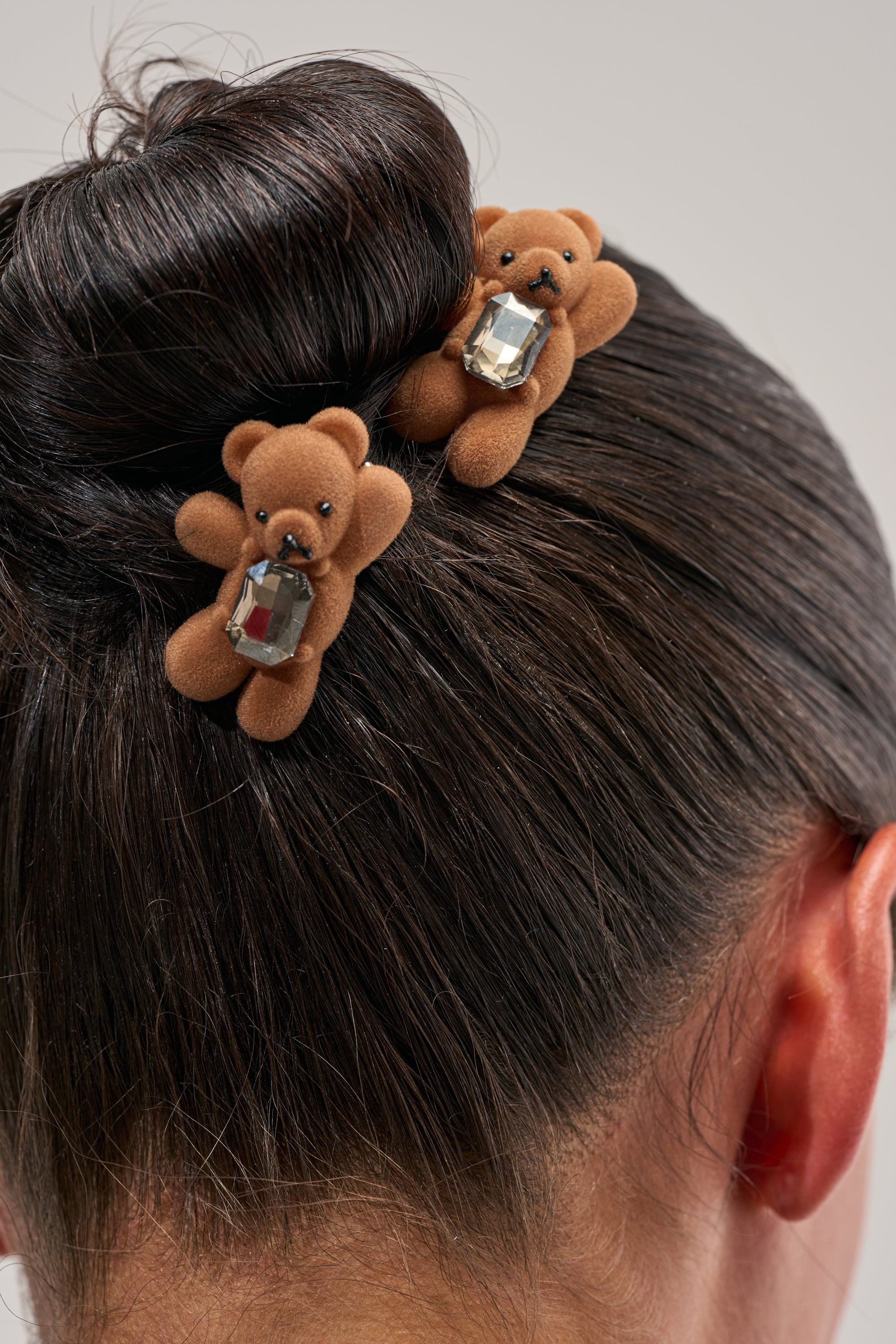 A close-up of dark hair styled in a bun, adorned with the BEARY CUTE HAIR CLIP SET—two brown teddy bear alligator clips, each embellished with a sparkling rhinestone gem.