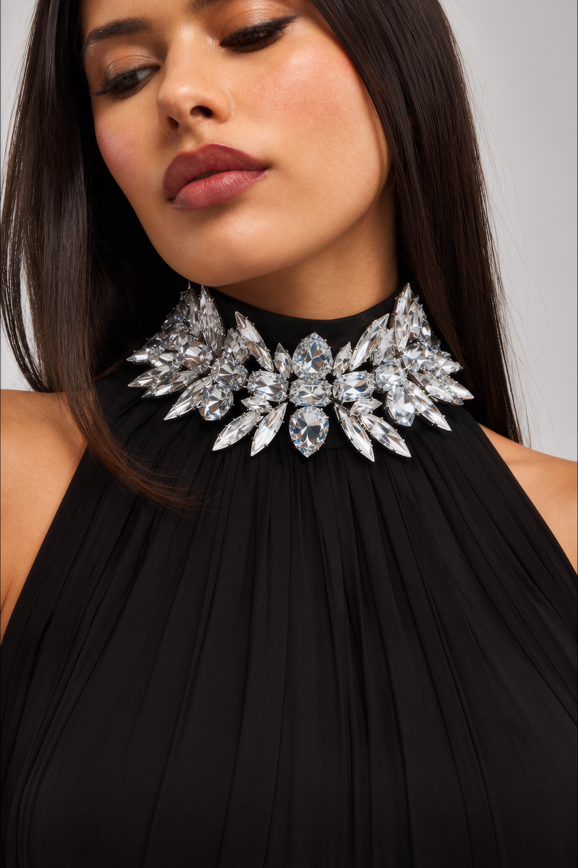 A woman wears the QUEEN OF IT ALL DIAMOND CHOKER—a bold, ornate crystal rhinestone statement necklace—paired with a high-neck black dress. With her straight dark hair and neutral makeup, she gazes downward, showcasing how this elegant choker makes the perfect sophisticated accessory for any birthday celebration.