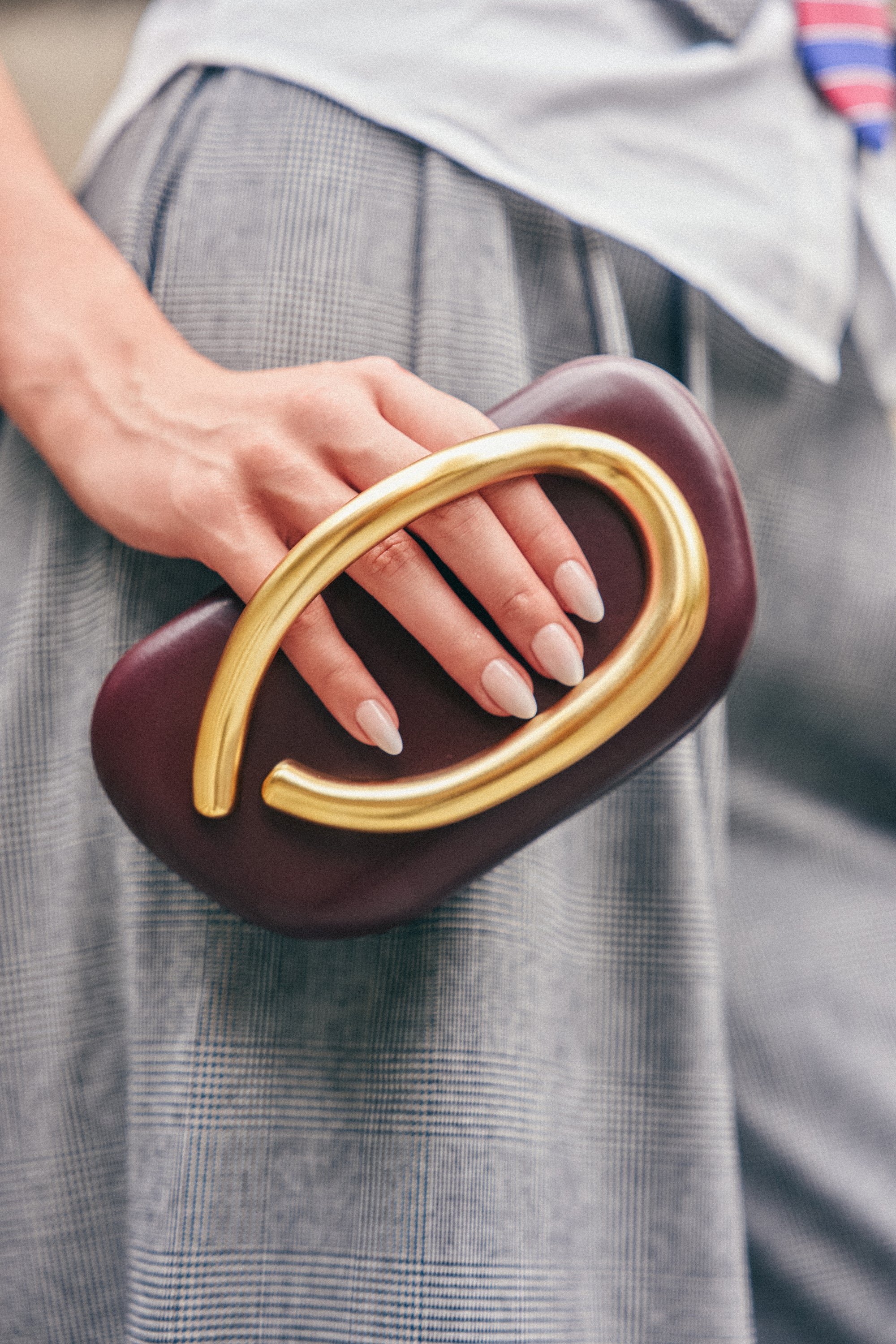 A person holding THE CUTEST BURGUNDY CLUTCH—a small, burgundy hardshell bag featuring a large, curved gold handle and elegant gold detailing, perfect as a wedding guest accessory—paired with a grey plaid skirt and light pink nail polish.