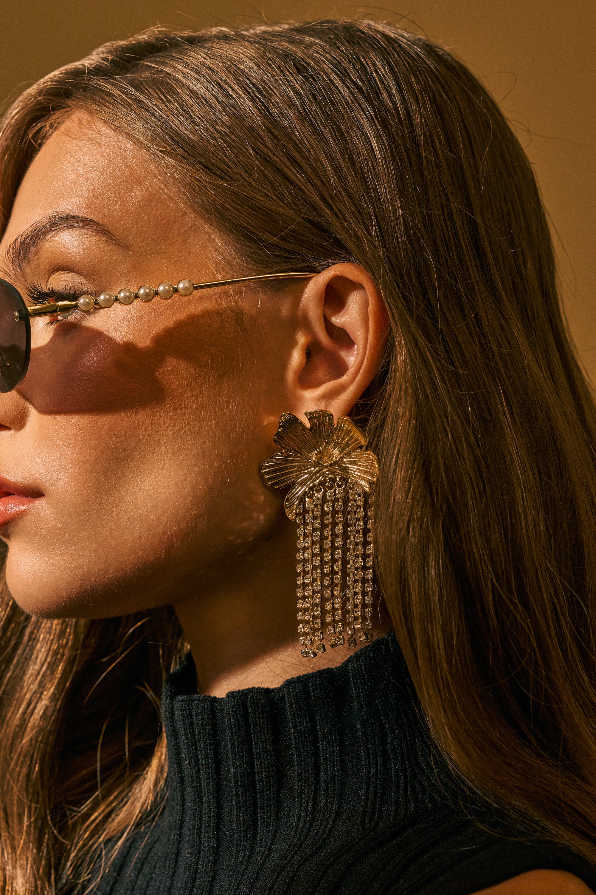 A woman with long brown hair wears pearl-embellished sunglasses and striking SHINE SOCIETY FLORAL DANGLE EARRINGS, paired with a black ribbed turtleneck top. The background is a warm beige color.