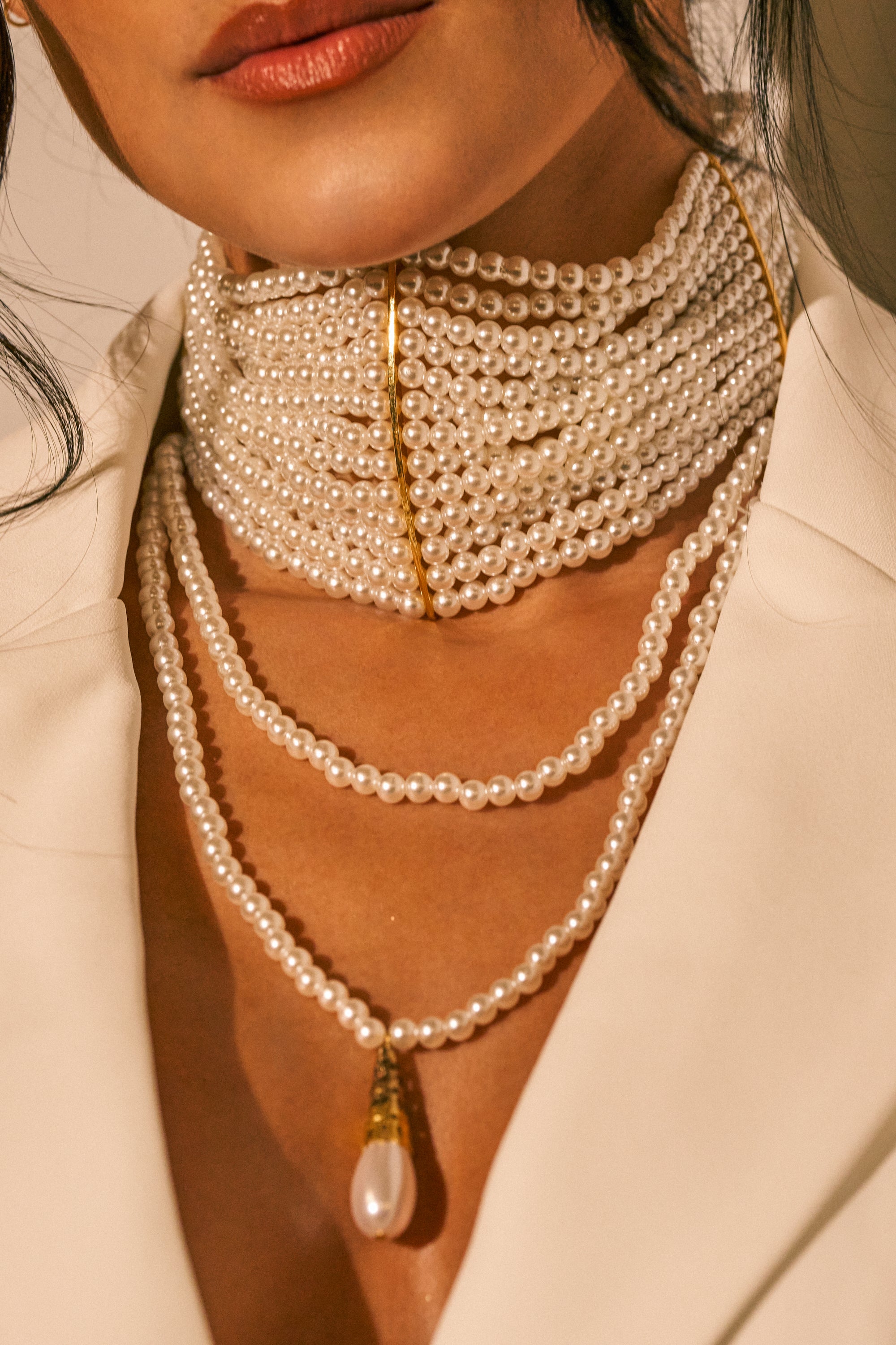 Wearing the PEARL GIRL LARIAT CHOKER—a faux pearl choker featuring gold accents and long layered strands, one adorned with a teardrop-shaped pendant—a woman pairs it with a cream-colored blazer for an effortlessly elegant look.