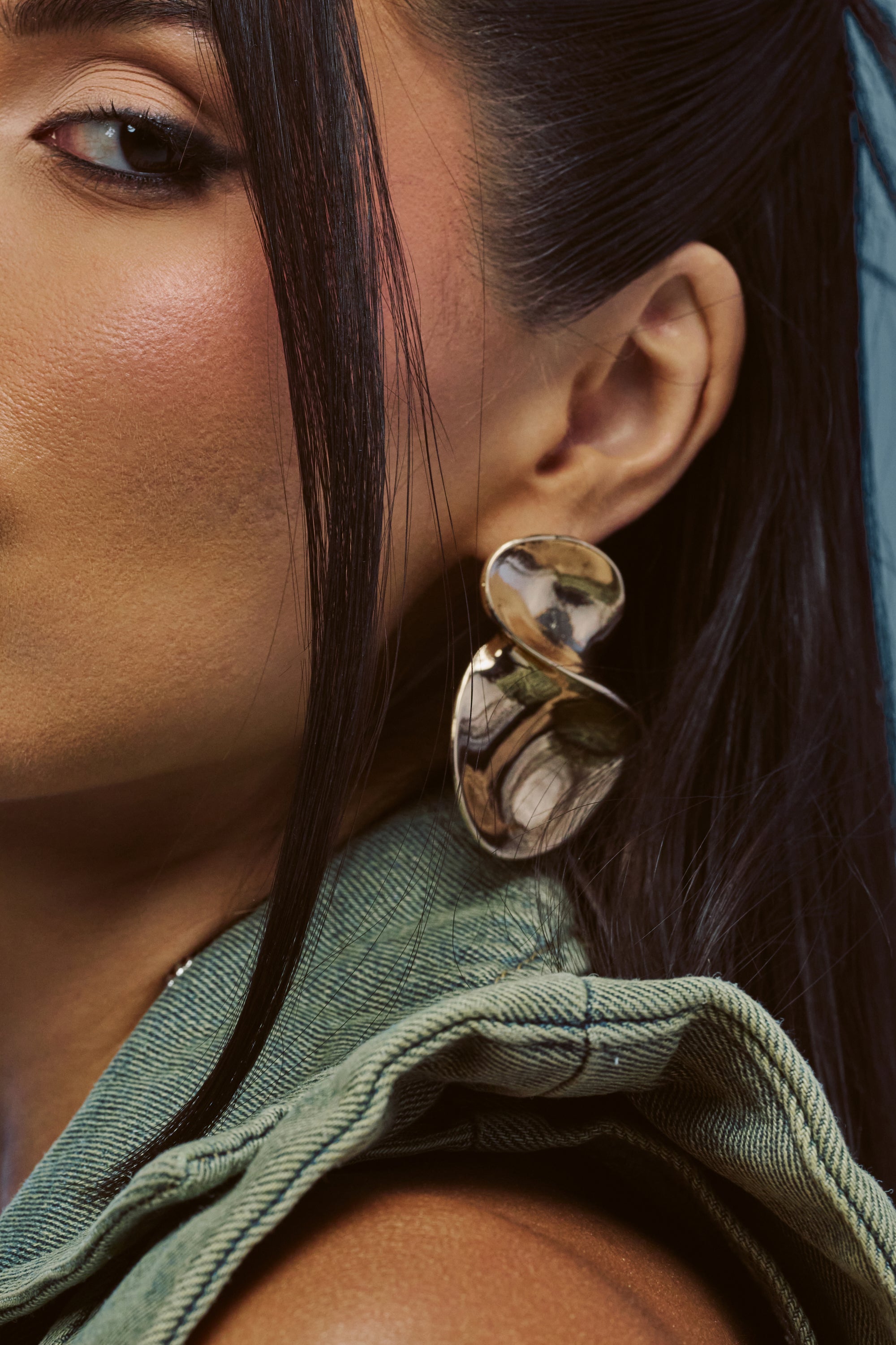 A close-up highlights a woman’s face and ear as she wears the SCULPTED FLORAL EARRINGS—bold, gleaming gold earrings with an abstract flower petal design, ideal for summer date nights. She has long dark hair, smooth skin, and is dressed in a green textured top with ruffled edging.