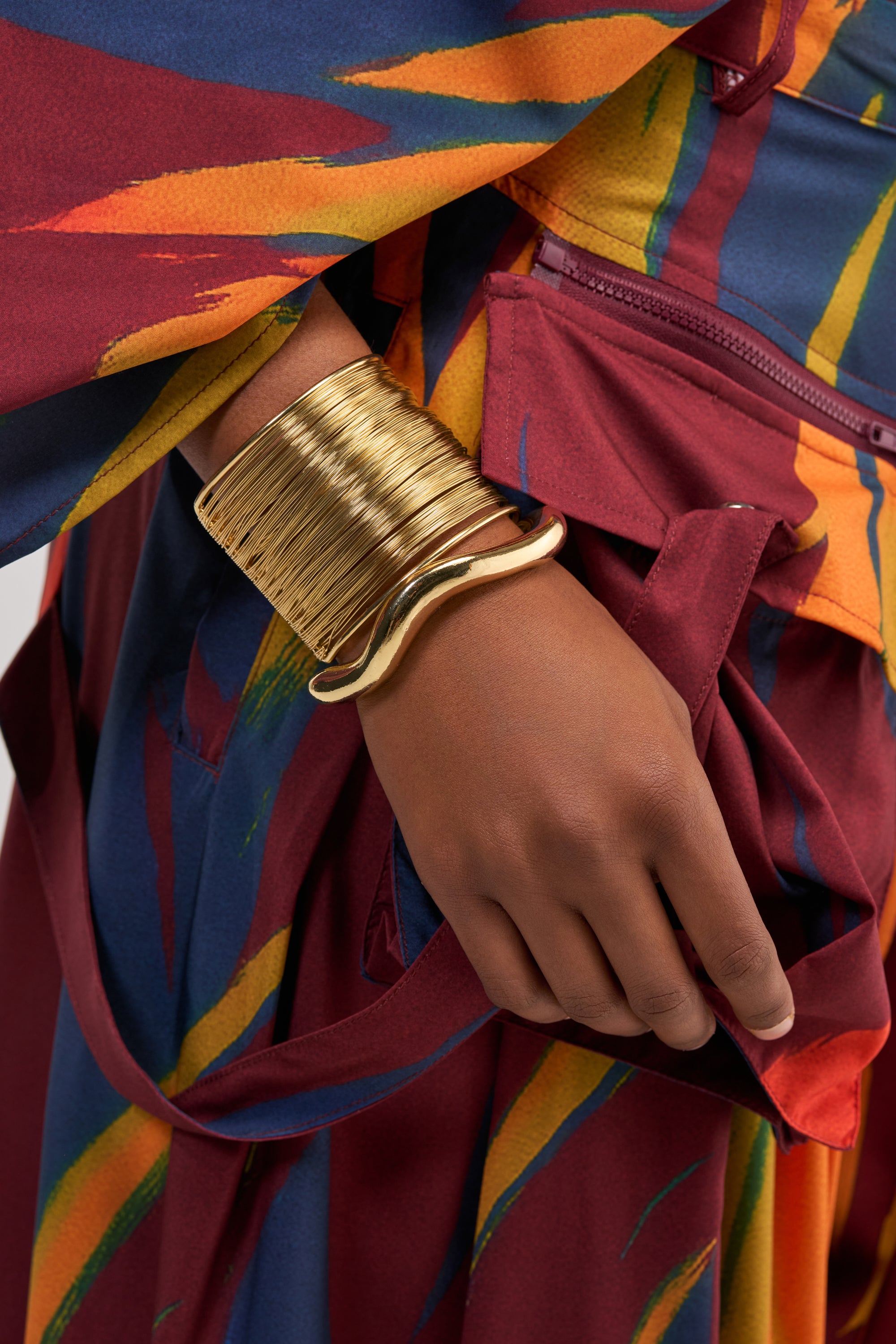 Wearing a vibrant, patterned outfit featuring maroon, yellow, and blue hues, this person accessorizes with the BOLD ENERGY CUFF SET—a striking set of wide, shiny gold bracelets that make a strong statement on their wrist.