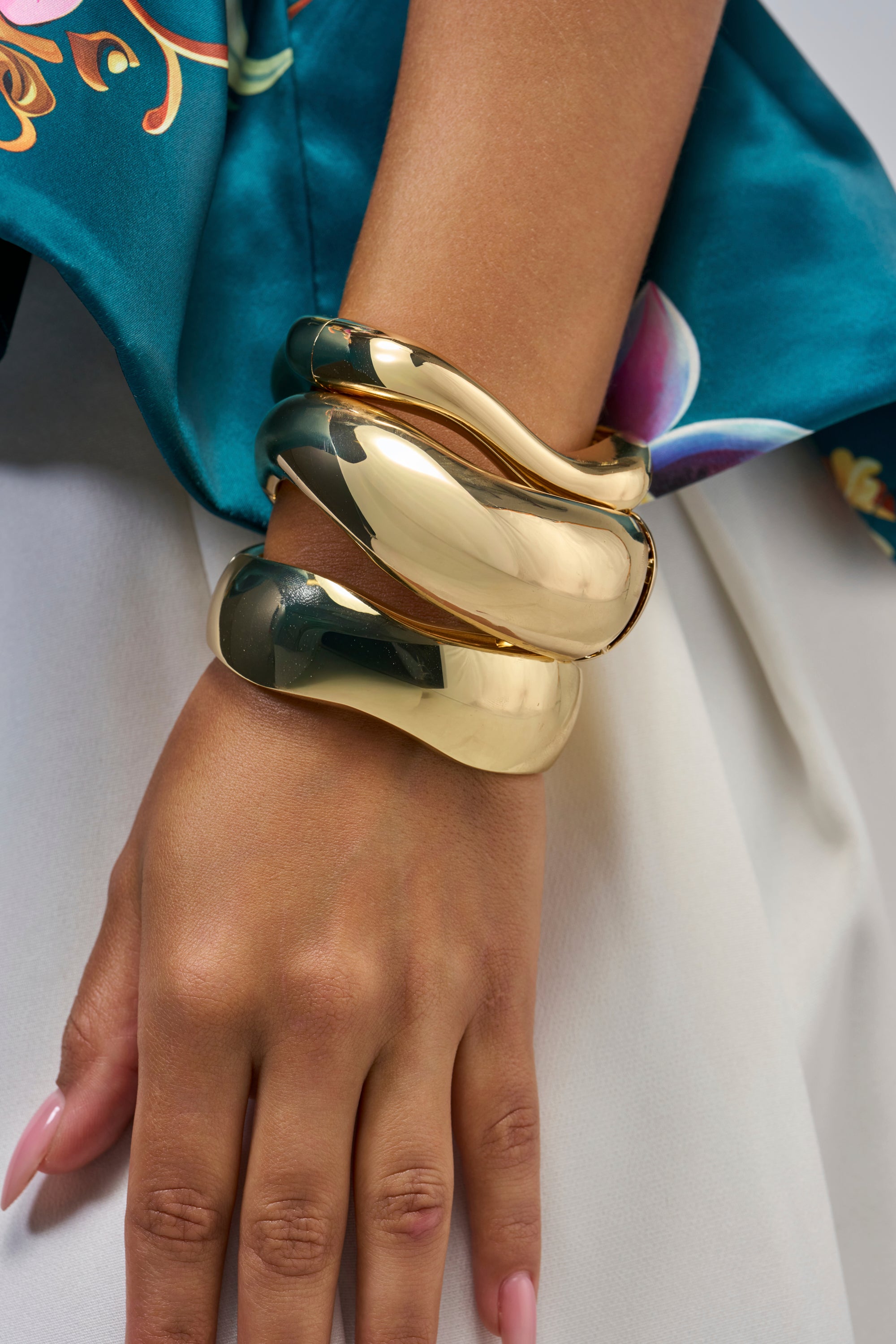 A hand adorned with two chunky, shiny gold bracelets from the GOLD ORGANIC BANGLE SET rests against a white skirt, with a teal floral-patterned fabric draped nearby.