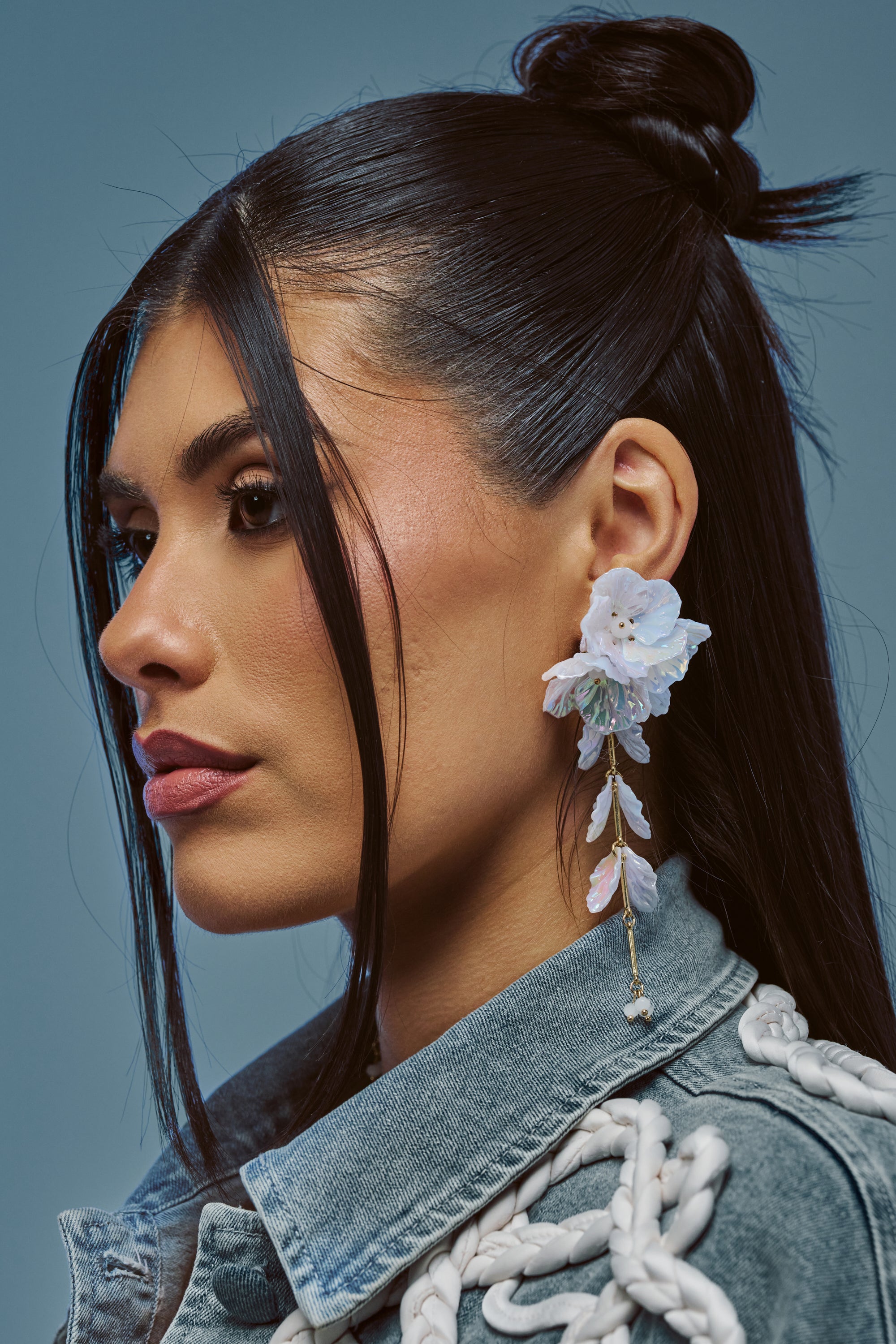 A woman in side profile with long dark hair styled half-up wears the IRIDESCENT FLORAL EARRINGS and a denim jacket with white rope details, set against a plain blue background.