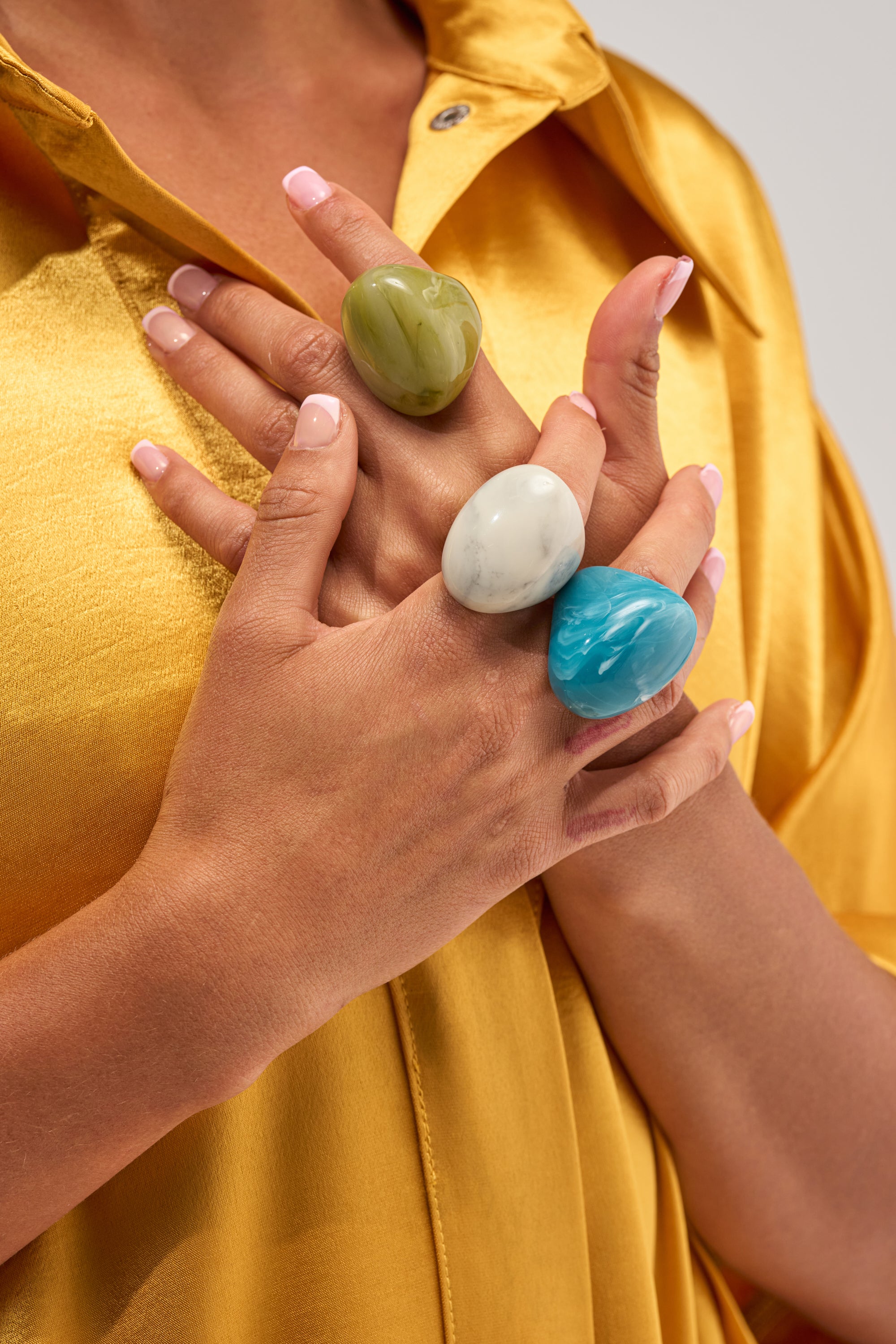Someone wearing a yellow shirt displays their hands adorned with the MULTI STACK RESIN RINGS in green, white, and blue, each chunky and colorful on their fingers. Their nails are neatly manicured with a light pink polish.