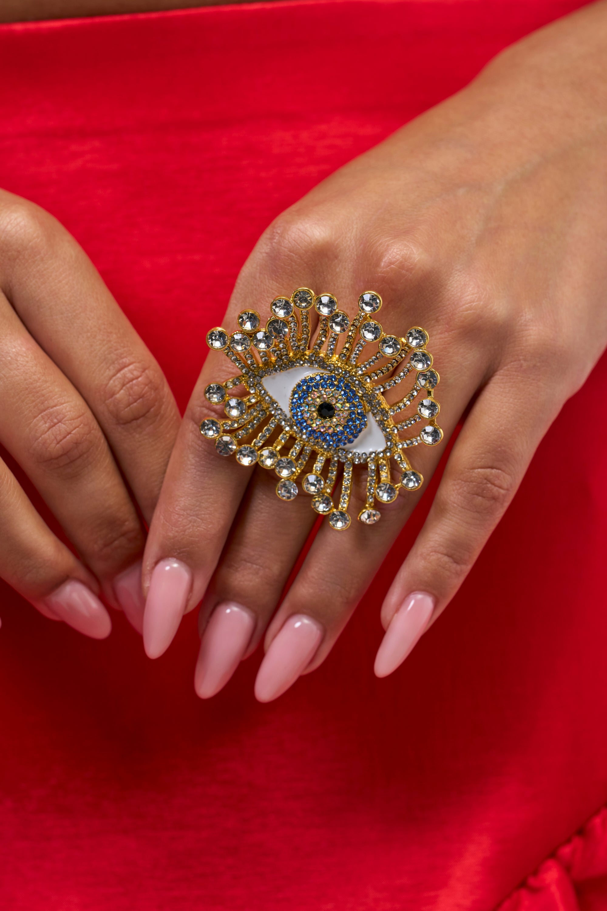 A hand with long, pink nails rests on red fabric, wearing the ICU STATEMENT RING—a large, gold ring shaped like an eye with blue, white, and gold detailing and sparkling gem accents.