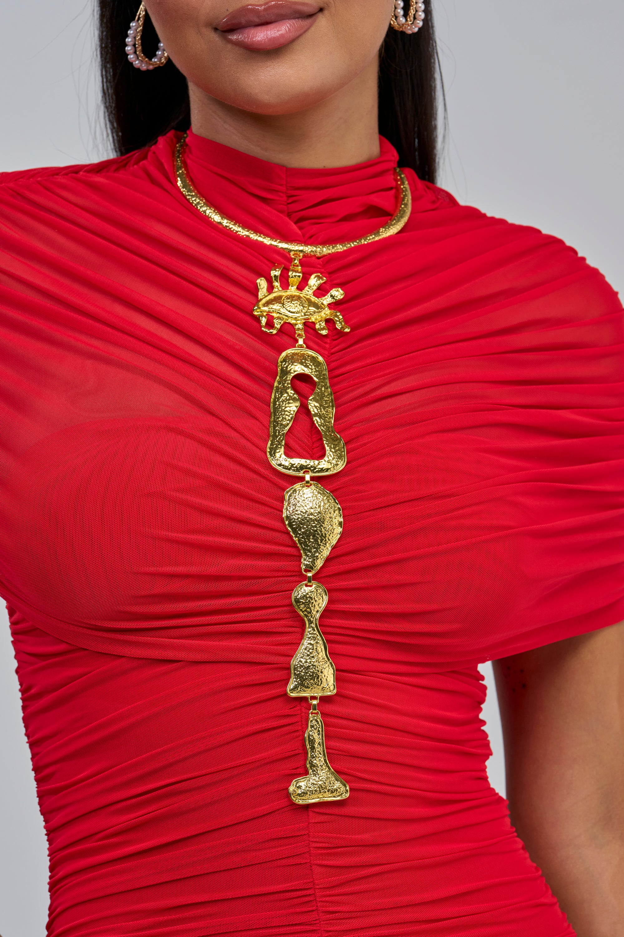 A woman wearing a bright red ruched dress and the DECONSTRUCTED DIVA CHOKER—a bold, sculptural hammered gold necklace with abstract shapes. Only her lower face, neck, and upper torso are visible.