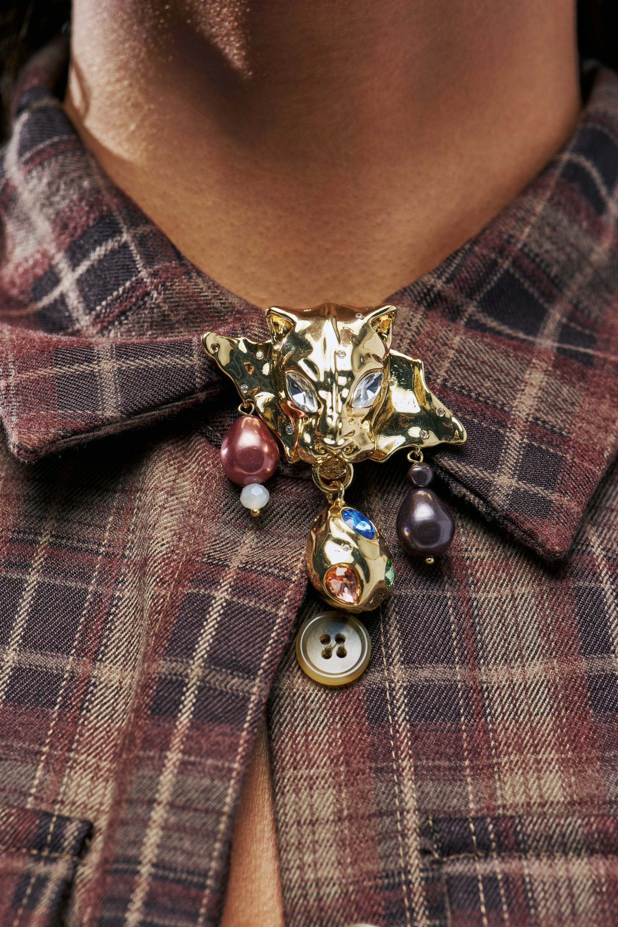 WILD THING BROOCH: A gold feline brooch shaped like a panther head, featuring dangling charms of pearls and faceted gemstones pinned to a plaid collared shirt.