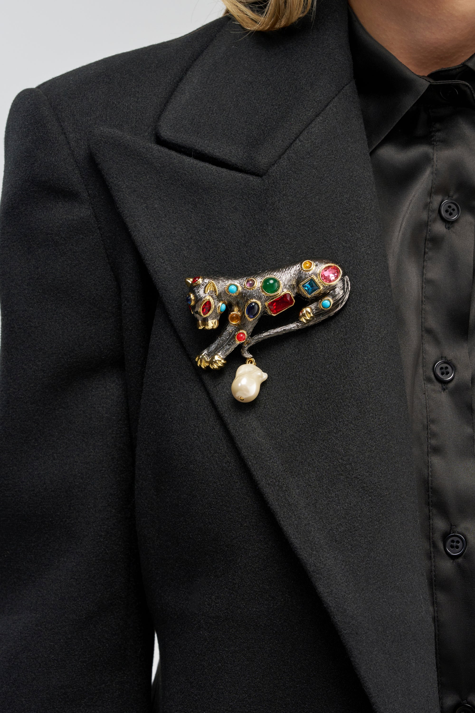 Wearing a black blazer and shirt, the person adds a bold statement to their workwear ensemble with the FERAL FANTASY BROOCH—a rhinestone-embellished feline brooch adorned with colorful gems and a hanging pearl on the lapel.
