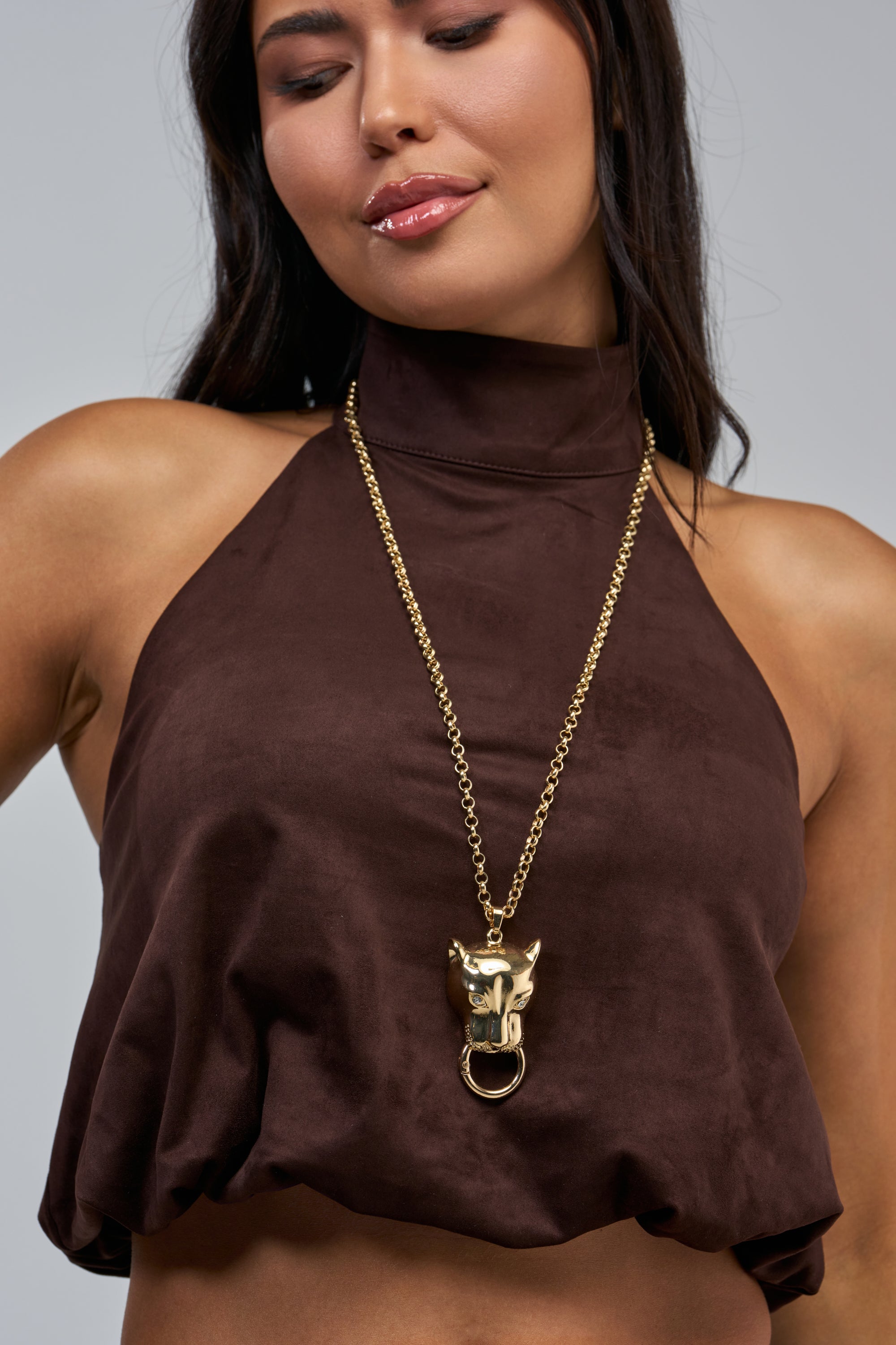 A woman in a brown halter top with a high neckline, wearing the NIGHT PROWLER NECKLACE—a gold statement piece featuring a bold panther head pendant with a ring in its mouth.
