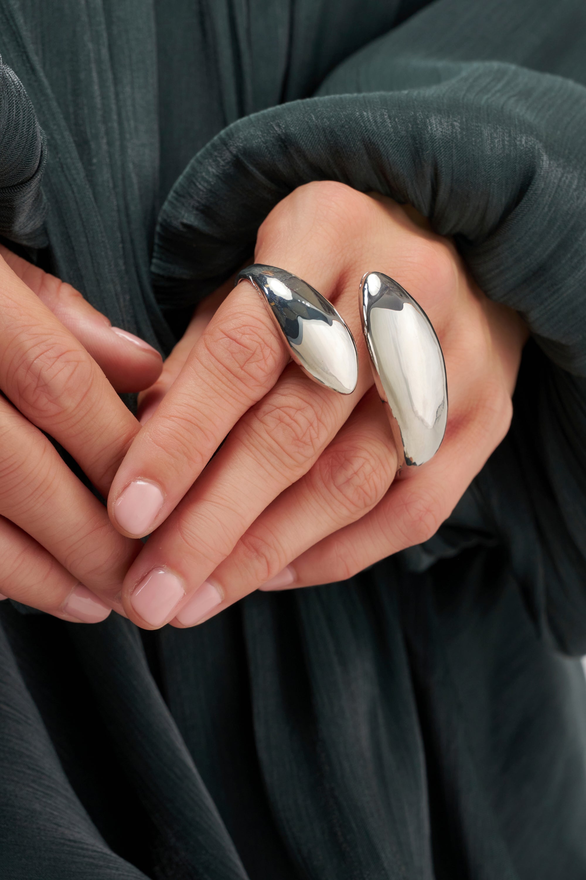 Two hands with neatly manicured nails, fingers gently touching, wear the FLUID FORM DOUBLE RING—a large, shiny silver piece that spans the index and middle fingers. This bold abstract accessory serves as an adjustable statement ring, perfectly complemented by the dark green textured sleeve in the background.