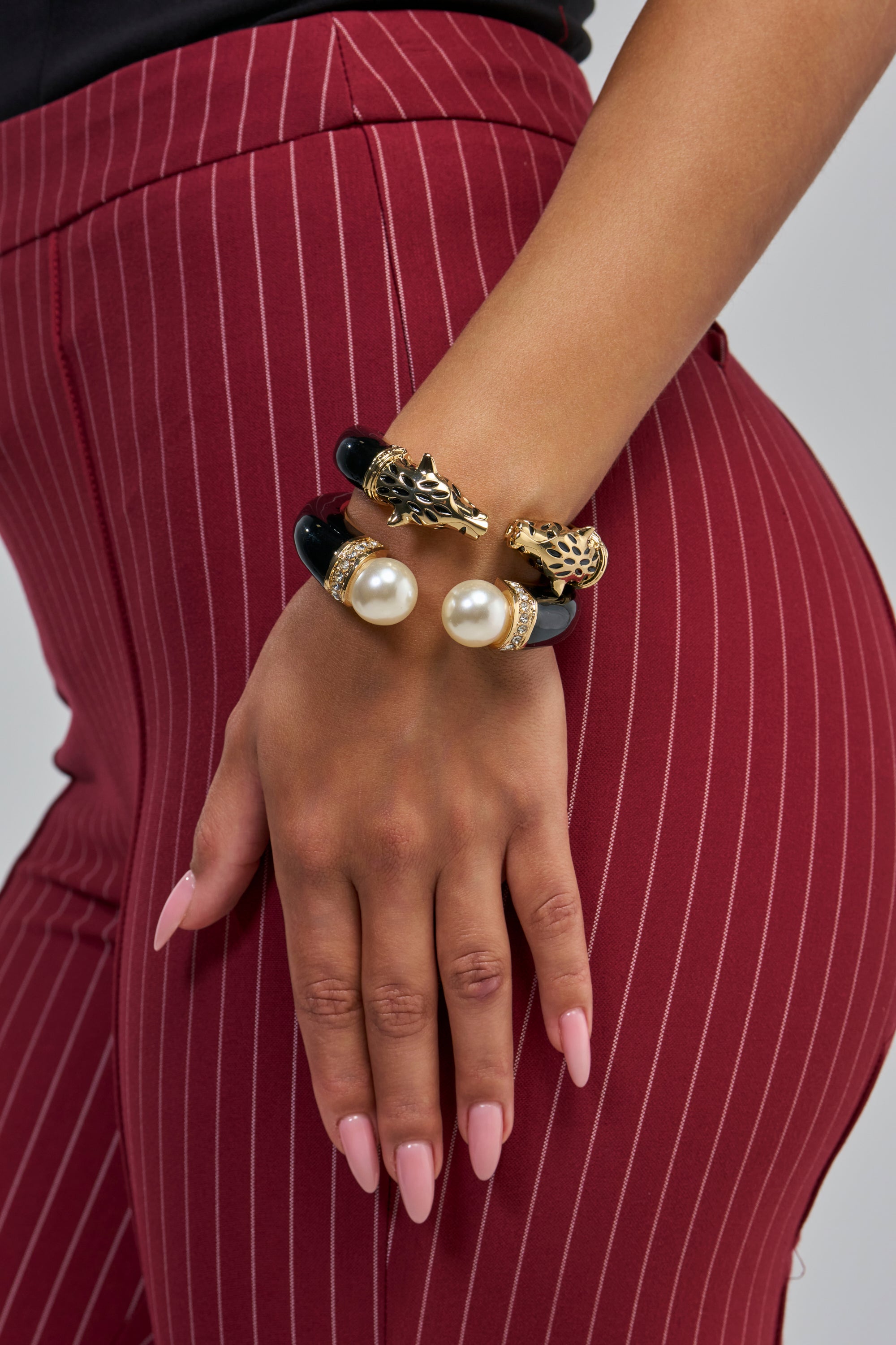 A hand with long, pale pink nails rests on the hip of someone in red pinstriped pants, showcasing the BLACK ENAMEL BRACELET SET adorned with feline motifs and large pearl-like accents.