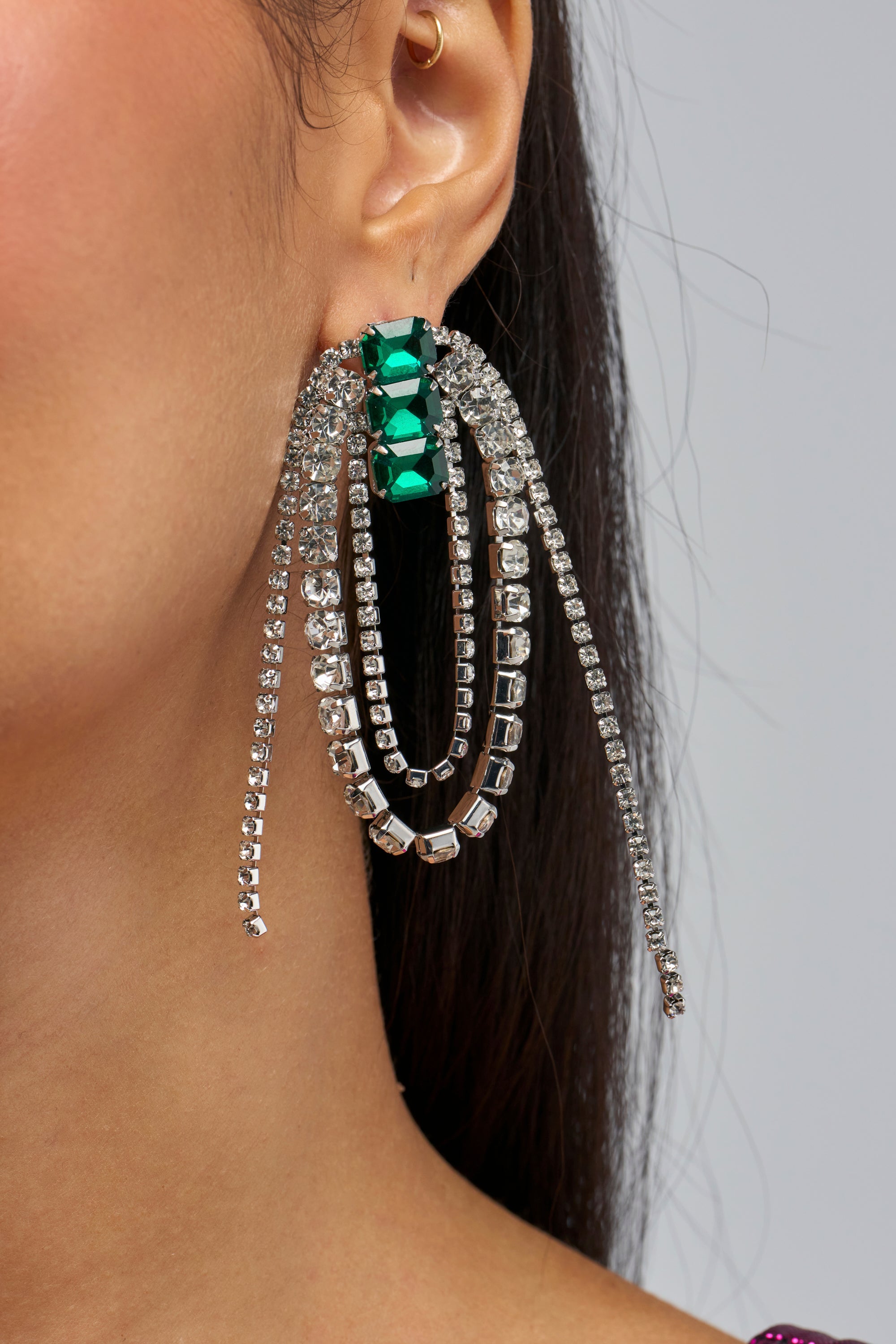 A close-up of a person’s ear wearing the EMERALD WATERFALL EARRINGS—large, sparkling rhinestone drop earrings featuring emerald accents and cascading silver rhinestone strands. The person also has a small gold hoop in the upper ear.