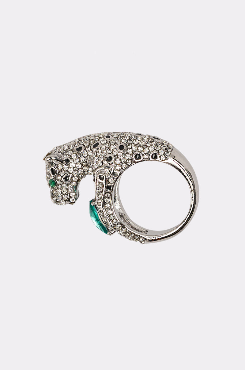 The LUXE JAG RING, inspired by a jaguar, features clear and black stones with emerald green eyes and a bold green gem below the head—a striking statement piece displayed on a plain white background. 0