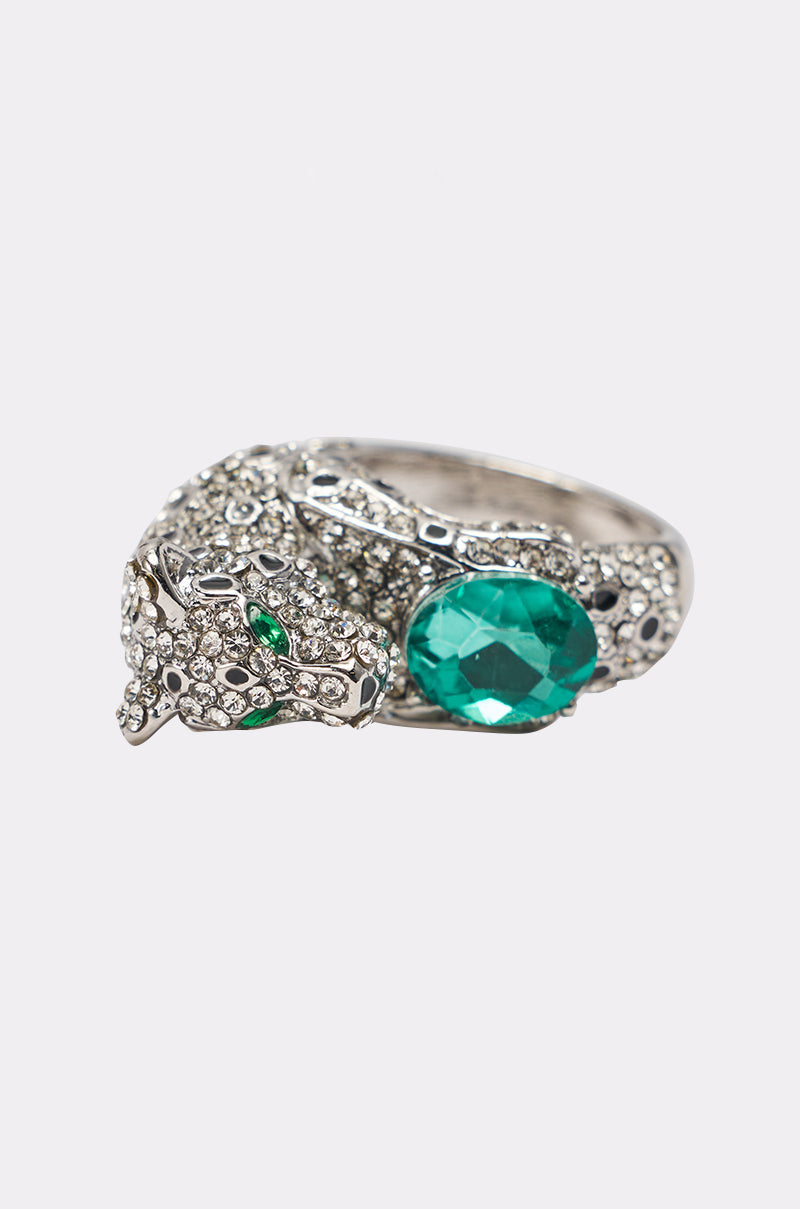 The LUXE JAG RING is a silver-tone panther-inspired band adorned with clear stones, featuring green gem eyes and a large oval green gemstone held between its paws—a perfect statement piece. 2
