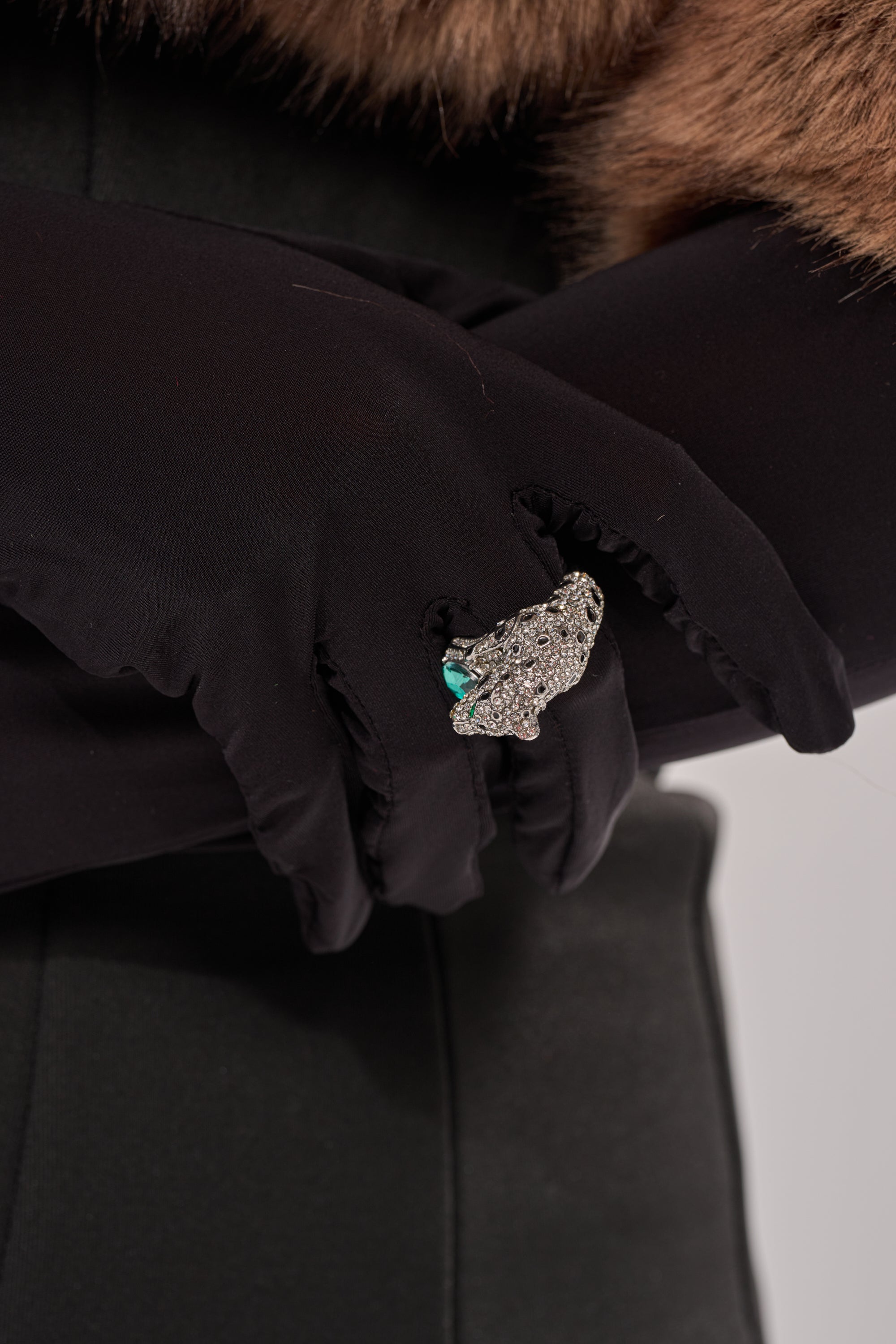 A person in black gloves and a dark coat with a brown fur collar crosses their arms, showcasing statement jewelry—the LUXE JAG RING, an ornate silver piece featuring a striking turquoise accent. 5
