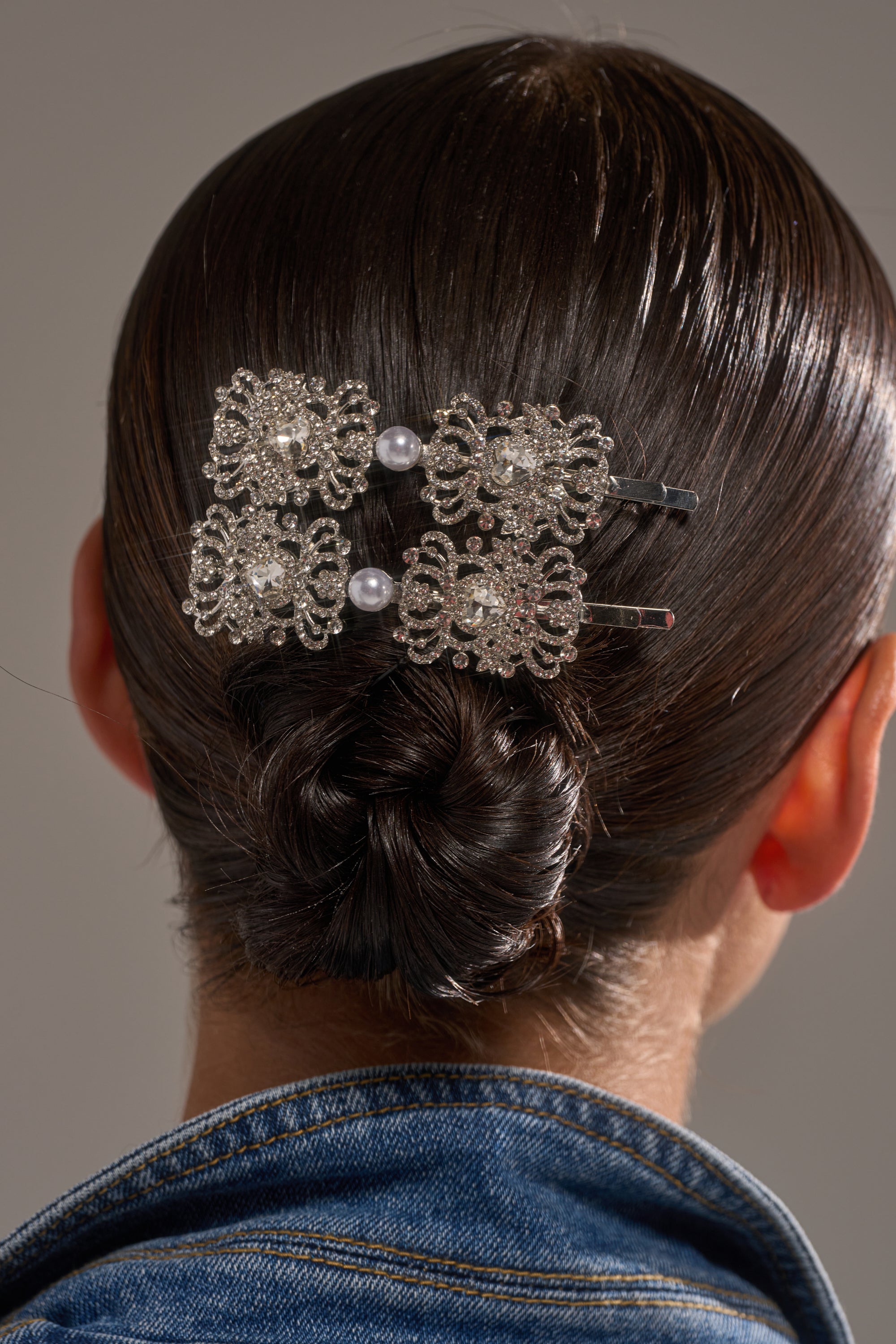A person with dark, neatly styled hair in a low bun wears the REGENCY HAIR CLIP SET, featuring ornate crystals and pearls, paired with a blue denim jacket. 0