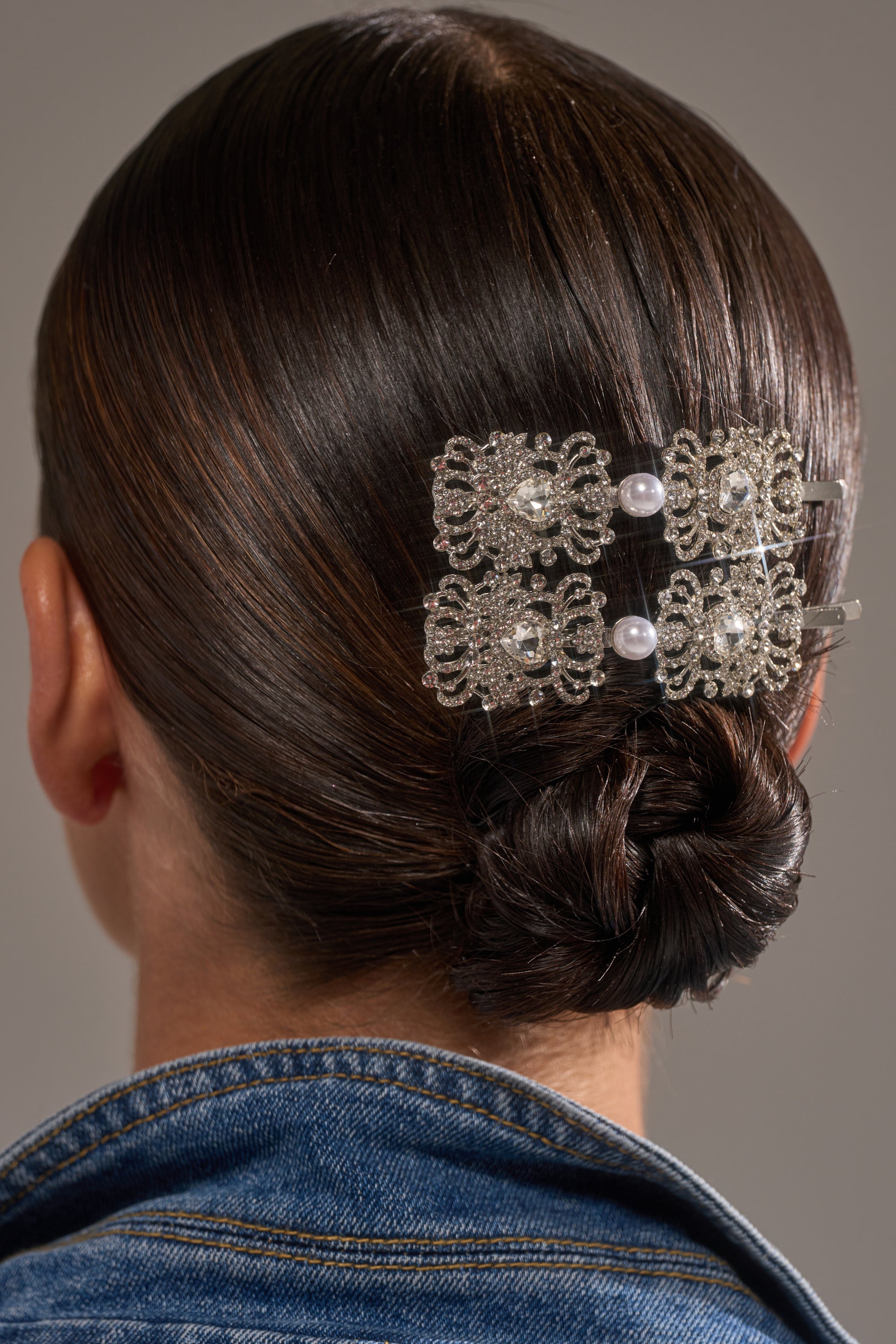 A close-up of dark brown hair styled in a low bun with the REGENCY HAIR CLIP SET—a vintage-inspired silver clip with pearls—paired with a blue denim jacket. 1