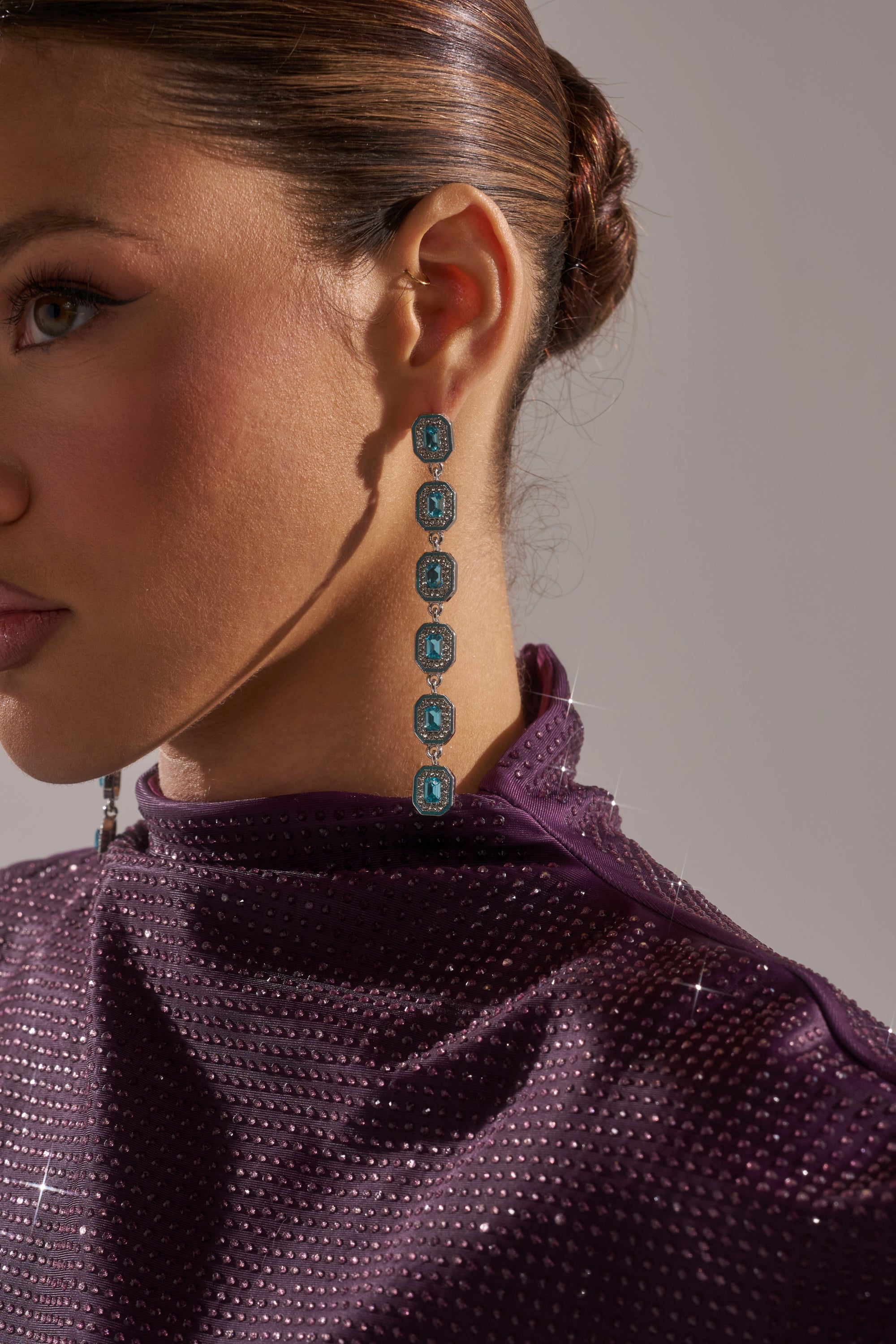 Close-up of a woman wearing the TOO RICH EARRING—emerald-cut aqua blue gems bordered with rhinestones. Her dark hair is in a neat bun, and she's dressed in a sparkling purple turtleneck top. 1