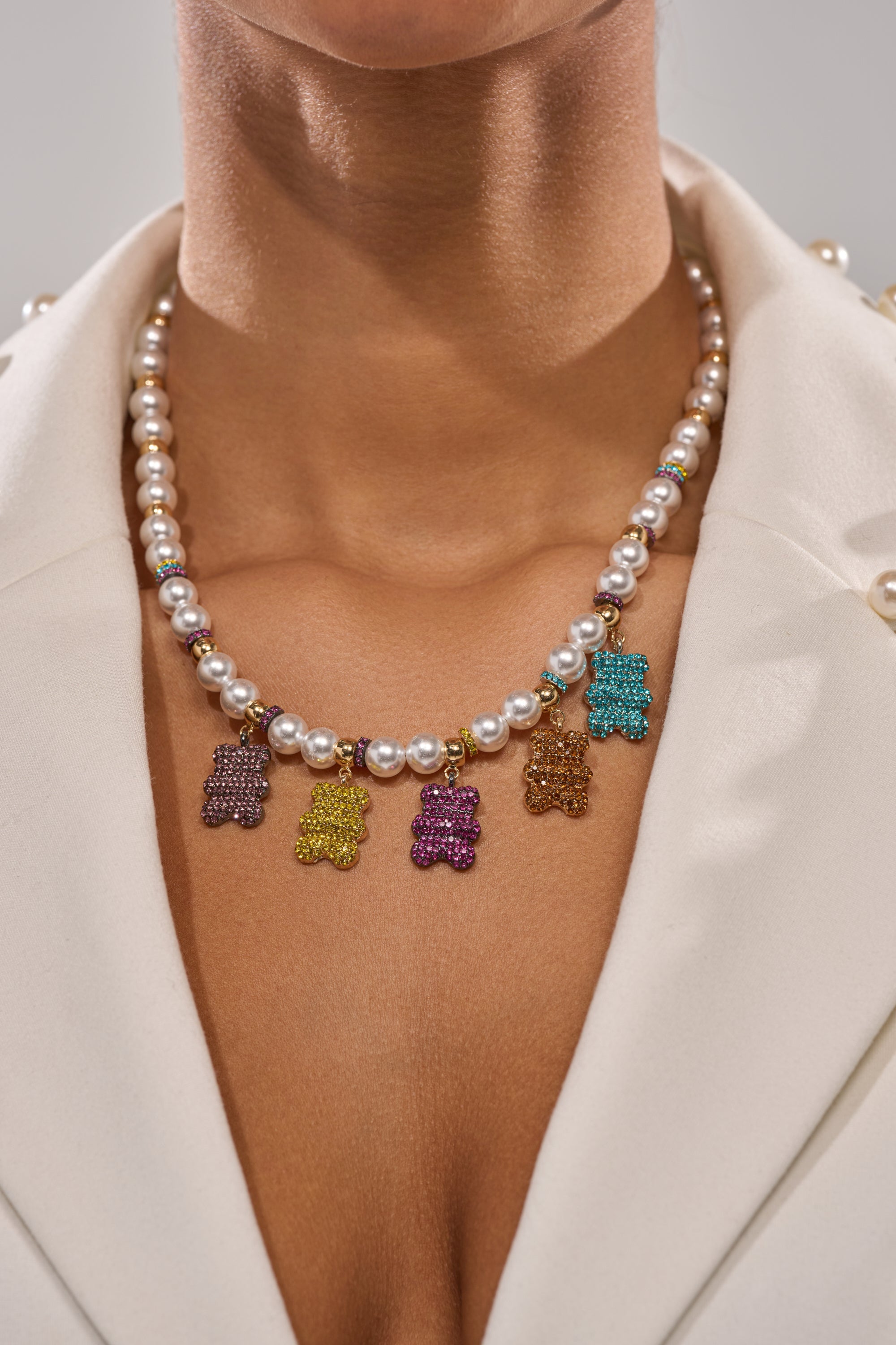 A person in a white blazer wears the LUXE BEAR CHARM NECKLACE, a faux pearl accessory featuring colorful gummy bear pendants in purple, yellow, pink, gold, and turquoise. The image highlights the neck and upper chest. 2