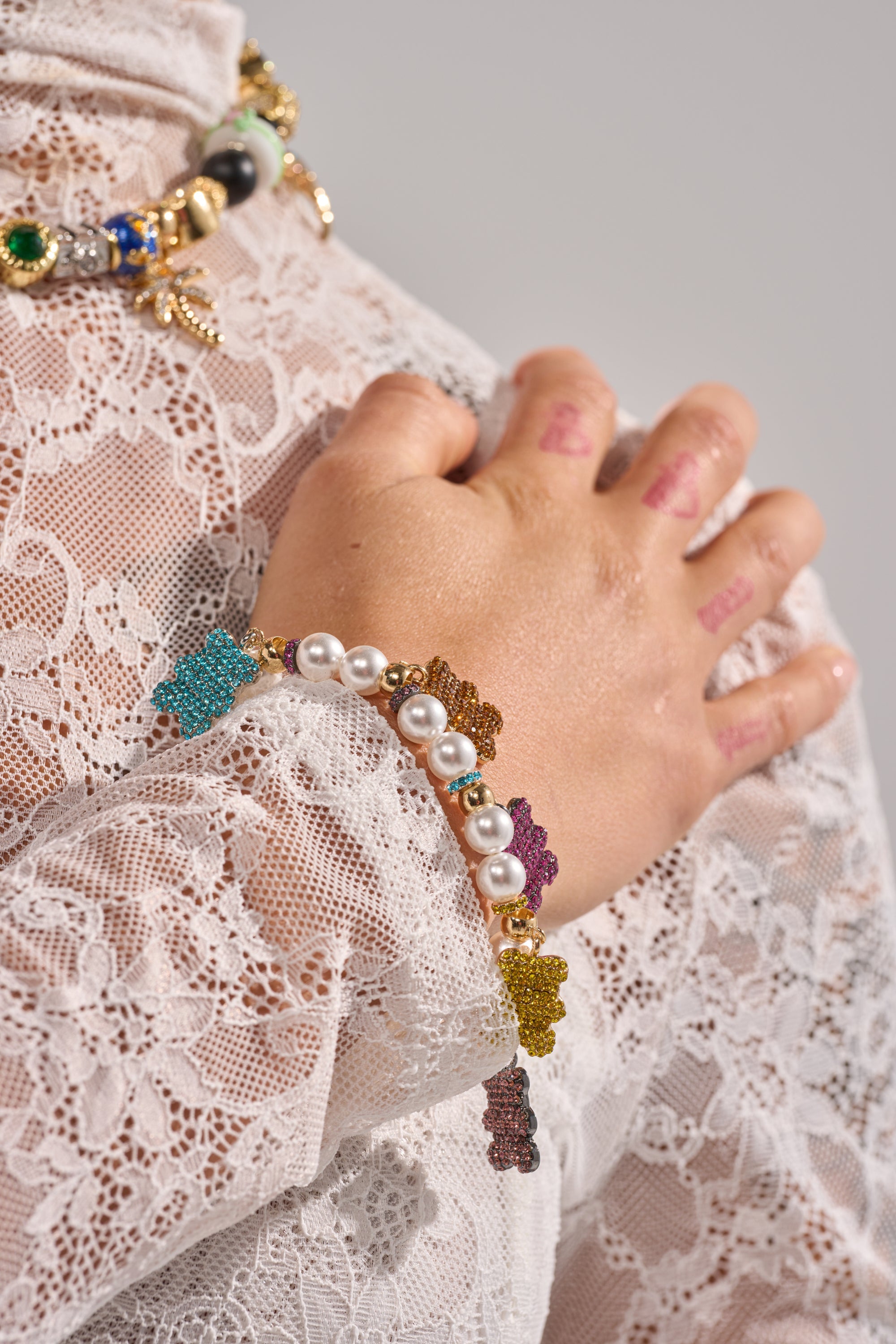 A hand with "THANK YOU" stamped on the fingers rests on a lacy white sleeve, adorned with the LUXE BEAR CHARM BRACELET—faux pearls and colorful bear charms—and an ornate green and gold rhinestone necklace. 1