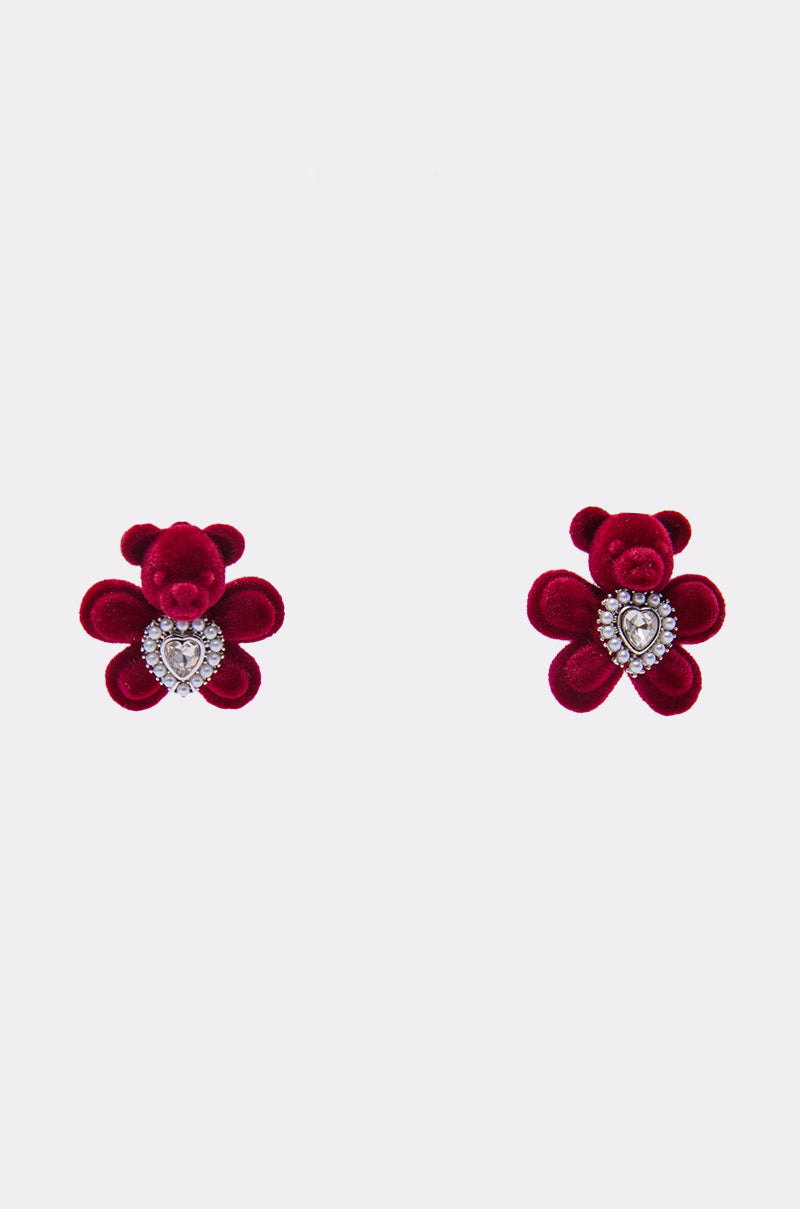 The HUG ME EARRING features two red velvet teddy bear studs with sparkling rhinestone heart details, adding a playful touch; these adorable earrings are beautifully displayed on a plain white background. 0