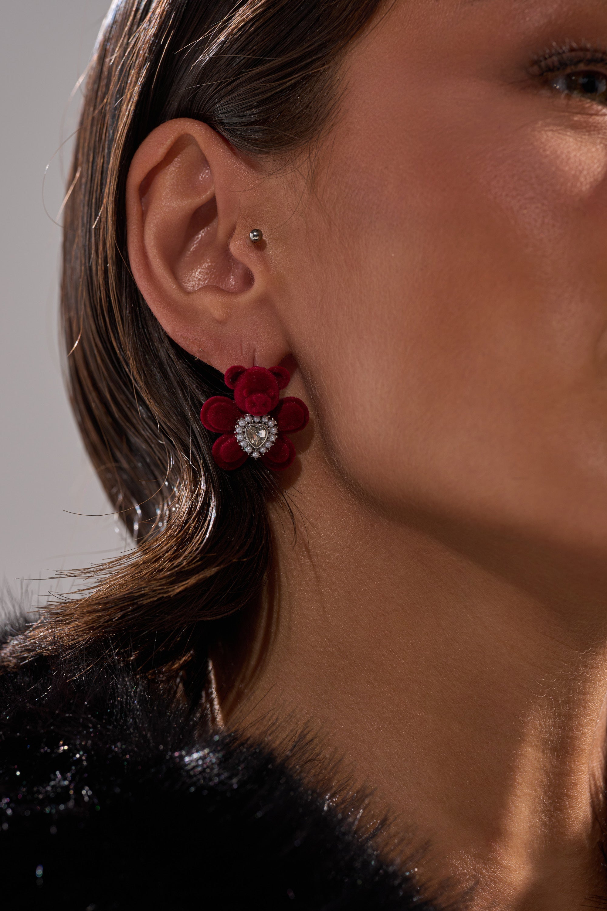 Close-up of a woman's ear wearing the HUG ME EARRING, a red flower-shaped piece with sparkling stones, paired with a velvet stud in her upper ear. She is dressed in a black, fuzzy garment. 1