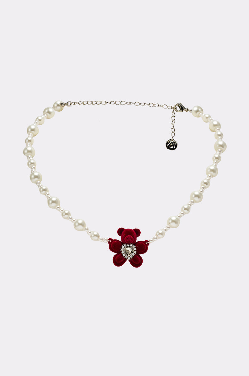 The HUG ME NECKLACE features a pearl chain with a red velvet bear pendant and heart-shaped gemstone center, shown on a light gray background. It includes an adjustable chain and a small round tag at the end.