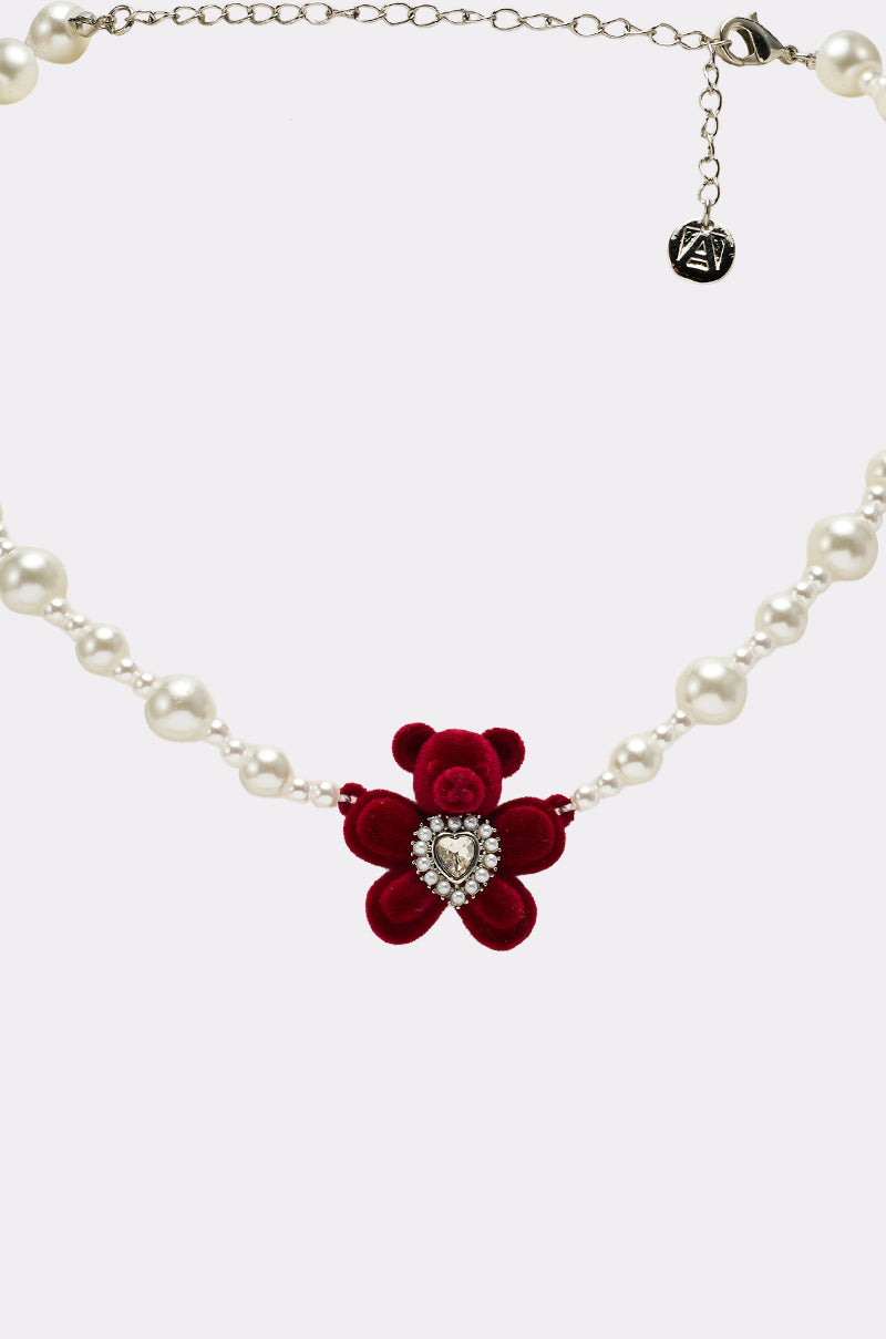 The HUG ME NECKLACE is a pearl necklace with a red velvet bear pendant featuring a heart gemstone on its chest and a small metal charm near the clasp.