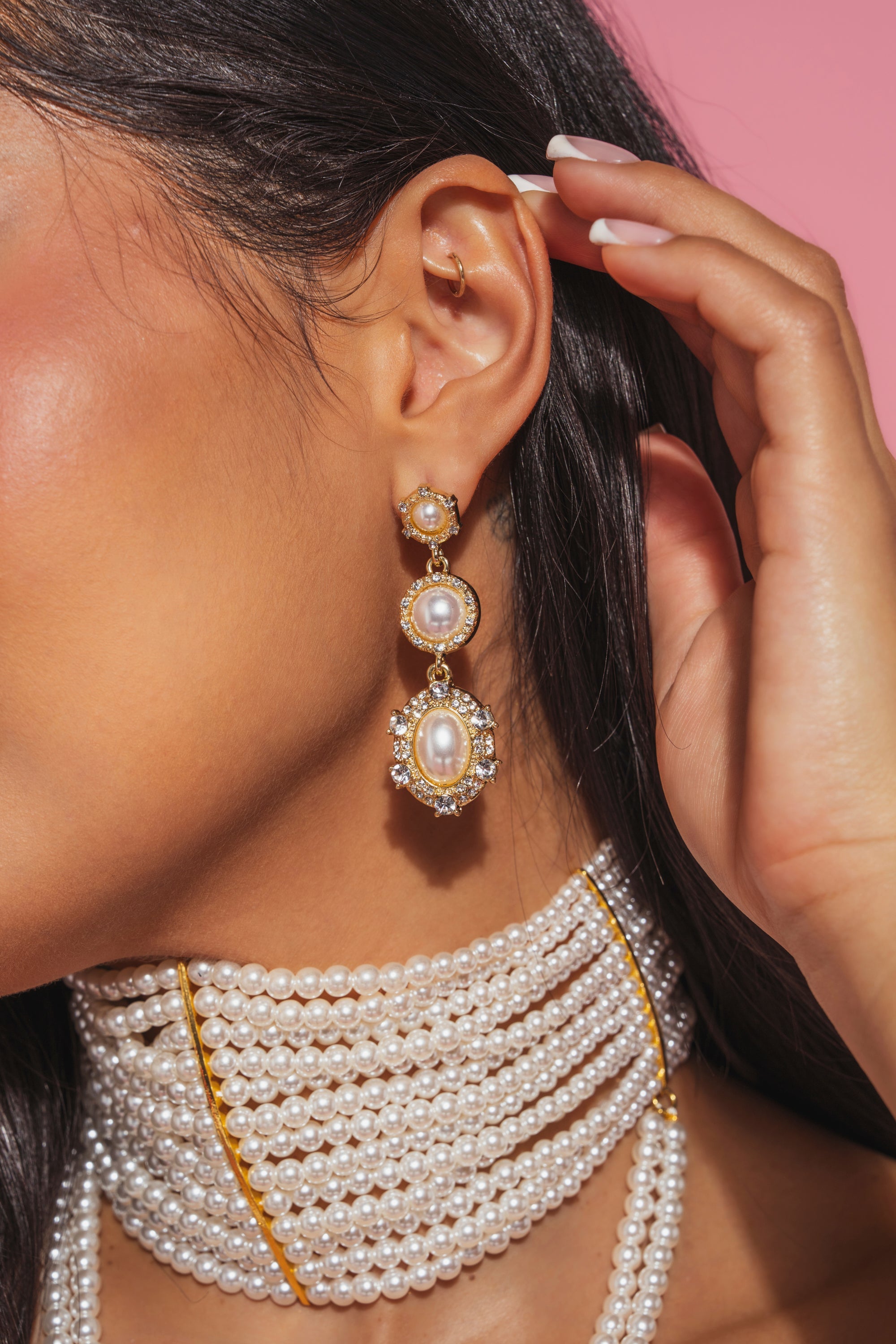 A woman with long dark hair wears the REGENCY EARRING—elegant, vintage-inspired jewelry—and a multi-strand pearl necklace. Her manicured hand touches her ear as she poses against a pink background. 0