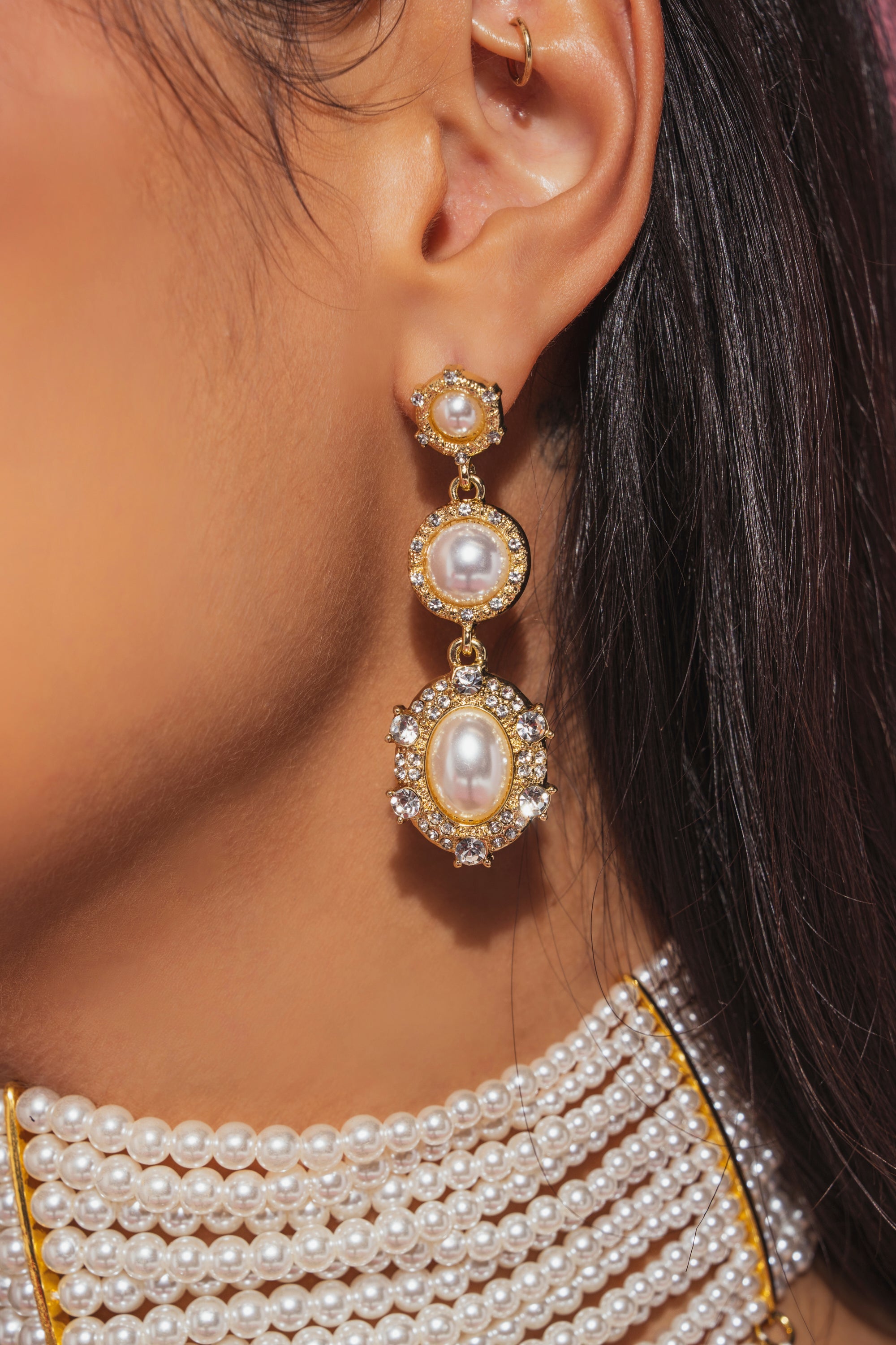 Close-up of a woman's ear and neck featuring the REGENCY EARRING—vintage-inspired pearl drop earrings with clear crystal accents—paired with a layered pearl necklace. The blurred background highlights the jewelry's exquisite details. 1