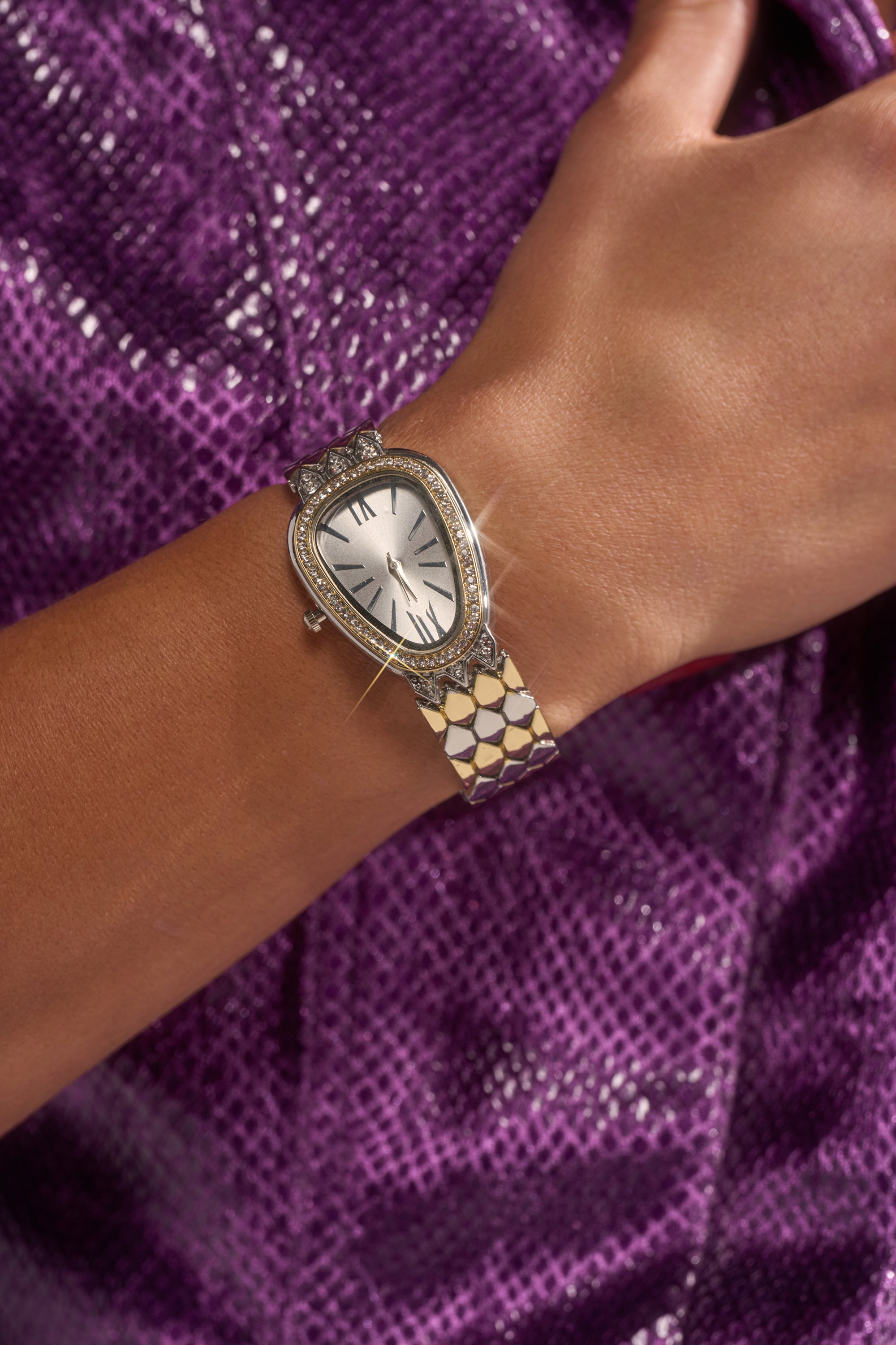 A close-up of a wrist adorned with the LUXE LIVING WATCH, featuring a sparkling, teardrop-shaped, diamond-studded snake design and an asymmetrical face, set against a backdrop of textured purple fabric.