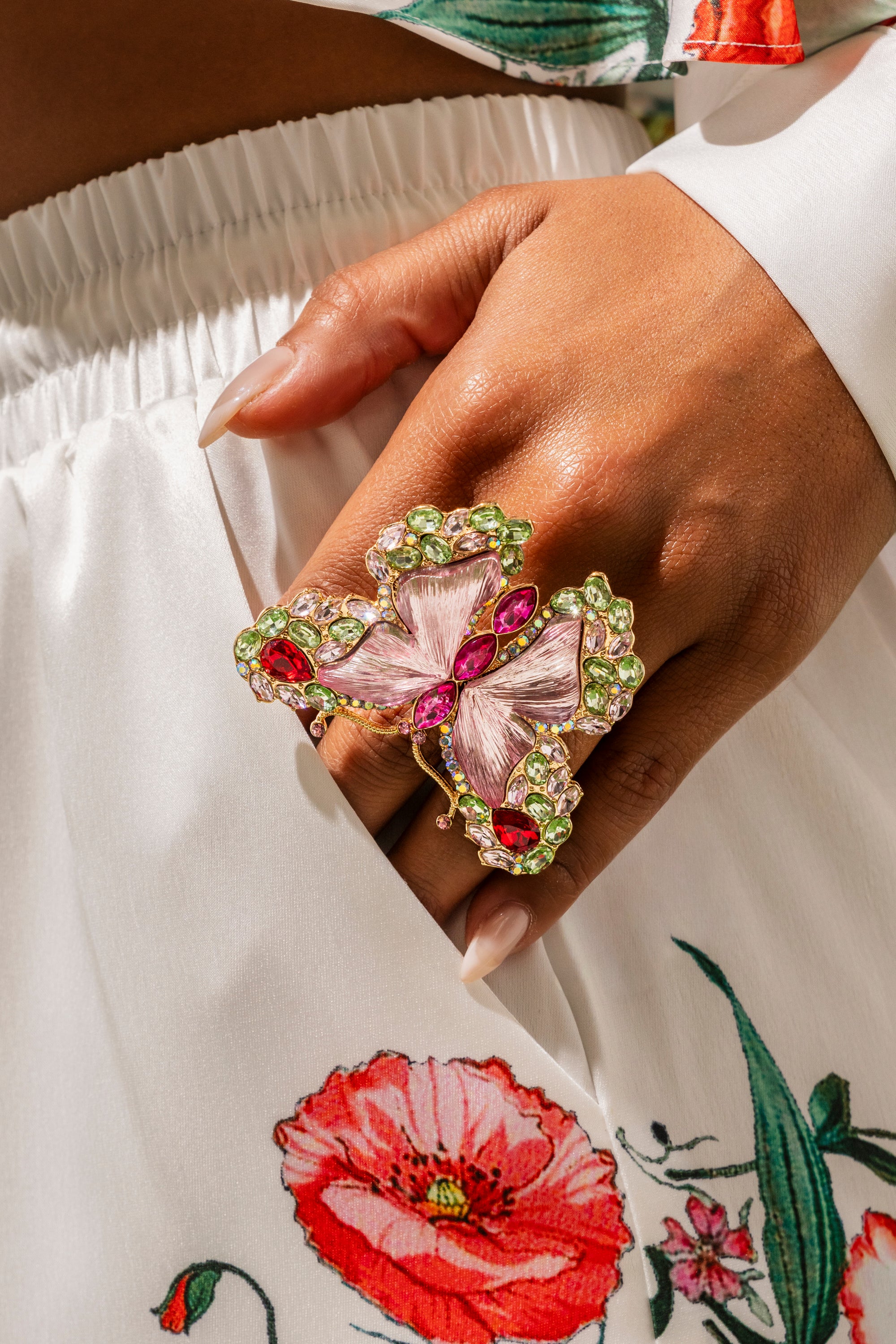 A hand with manicured nails showcases the SPREAD YOUR WINGS RING, a bold butterfly design featuring pink, green, and red gemstones, resting against white fabric with a red floral print. 1